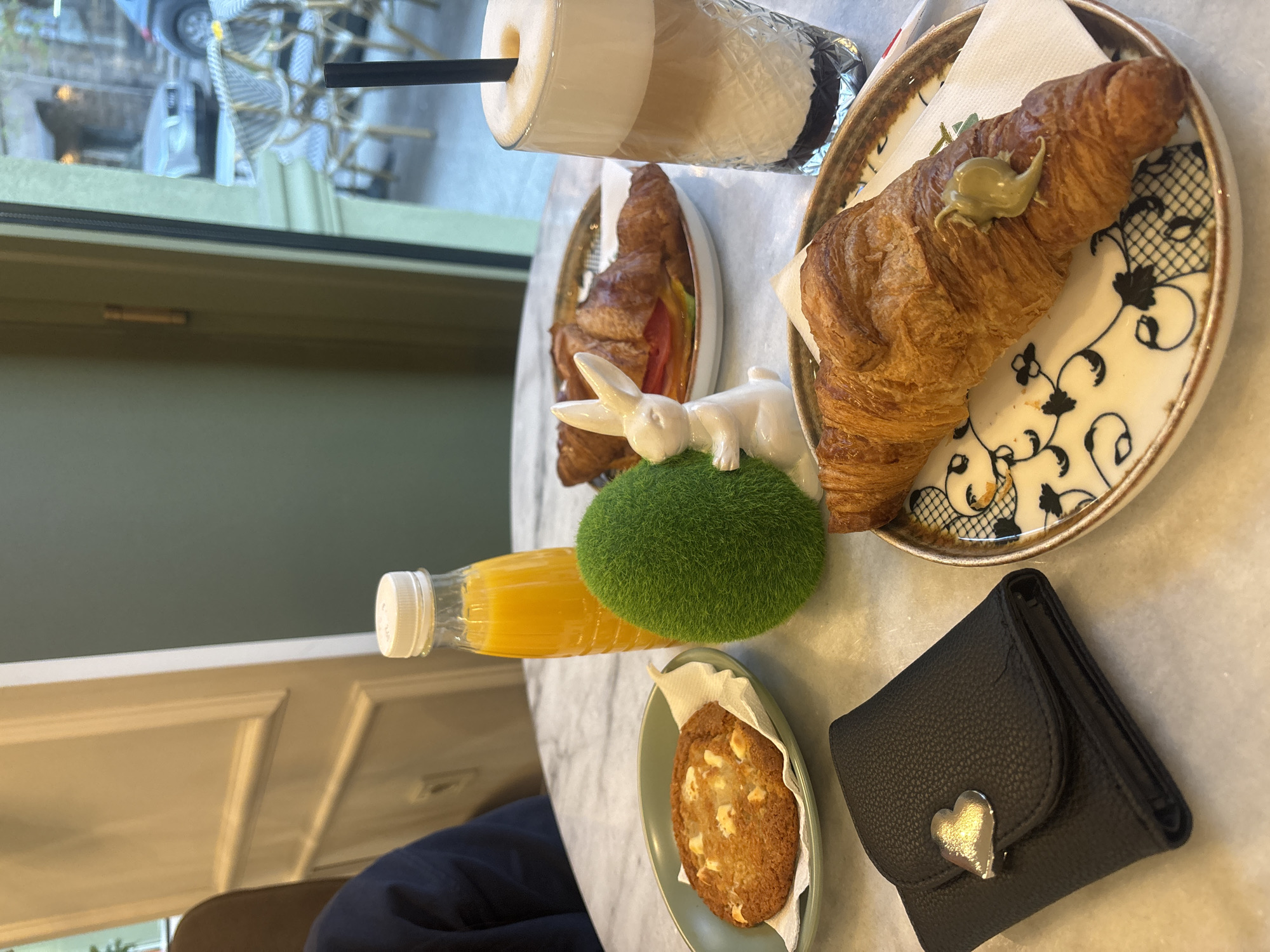 The image captures a breakfast or brunch scene set on a marble-top table, likely indoors. A glass of layered coffee with a black straw is positioned on the table. Nearby, there are two plates of croissants: one with a single croissant topped with a green paste, set on a decorative plate with black and white patterns, and another with a croissant sandwich featuring visible red tomato slices. A bottle of orange juice lies horizontally near a green, spherical object. A small cookie on a paper napkin is placed on a green plate. A black wallet, embellished with a heart-shaped metal detail, sits on the table's surface. A white rabbit figurine is placed on top of the green spherical object. Through a window, elements of an outdoor scene are visible, including a car and bistro-style chairs and tables. There is no verifiable information about the location of the image, Burgas, Bulgaria.