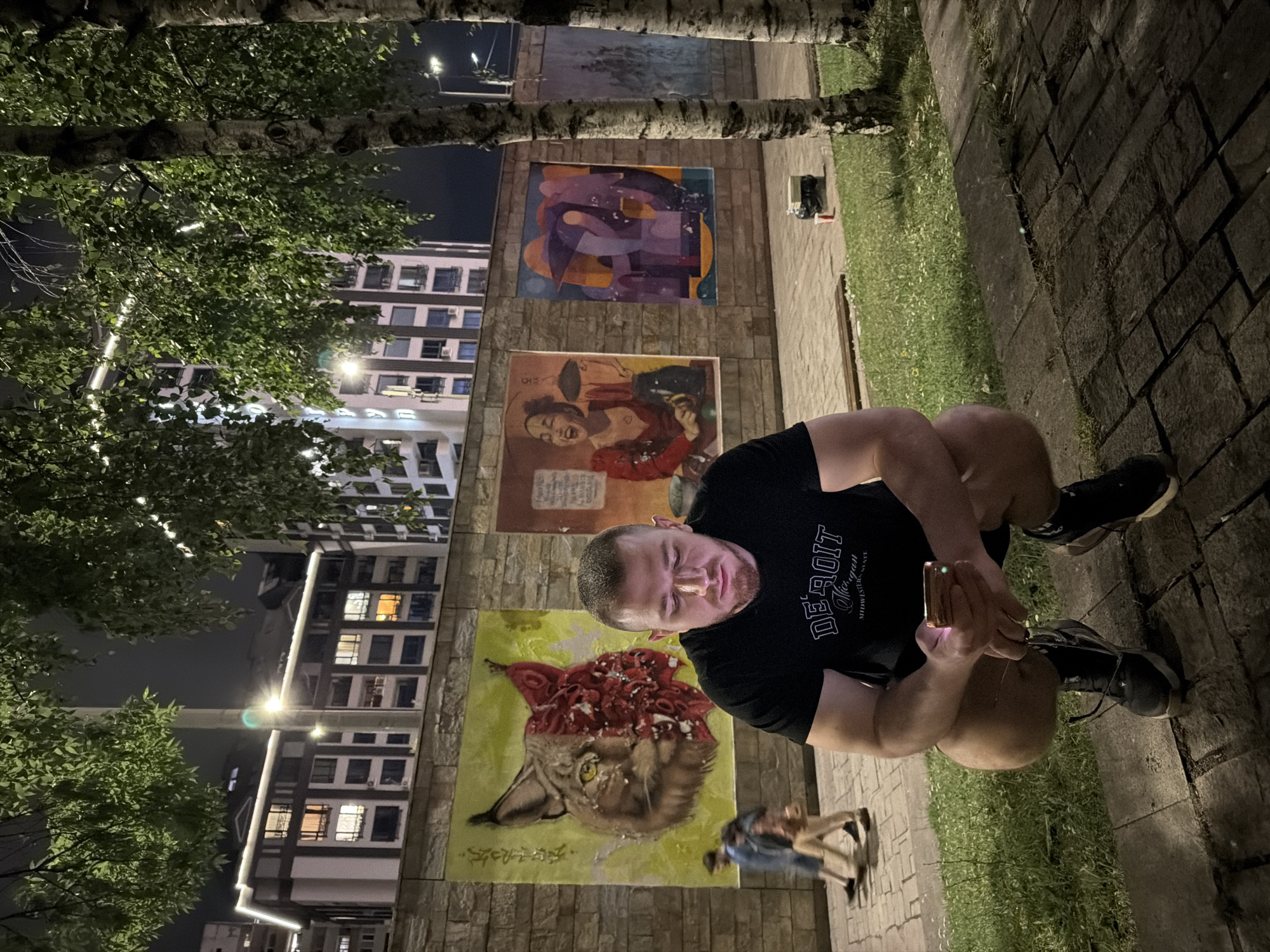A man is squatting on a cobblestone path in front of a brick wall adorned with three distinct murals, set against a backdrop of city buildings and trees at night. The man, wearing a black t-shirt and shorts, is looking down at his phone, which is illuminated and held in his hands. To his left, the first mural depicts the face of a lynx, half rendered in a realistic style and the other half in a more abstract, textured manner. The middle mural features a woman in a red dress, laughing and holding a black object. The third mural, on the right, displays abstract shapes in purple and orange hues. In the background, several multi-story buildings are visible, their windows lit up. Two people are walking in the distance, on the left side of the frame. The ground is a mix of grass and paving stones, with a few small trees casting shadows. The overall atmosphere is urban and nocturnal. The image does not show Sofia, Bulgaria.