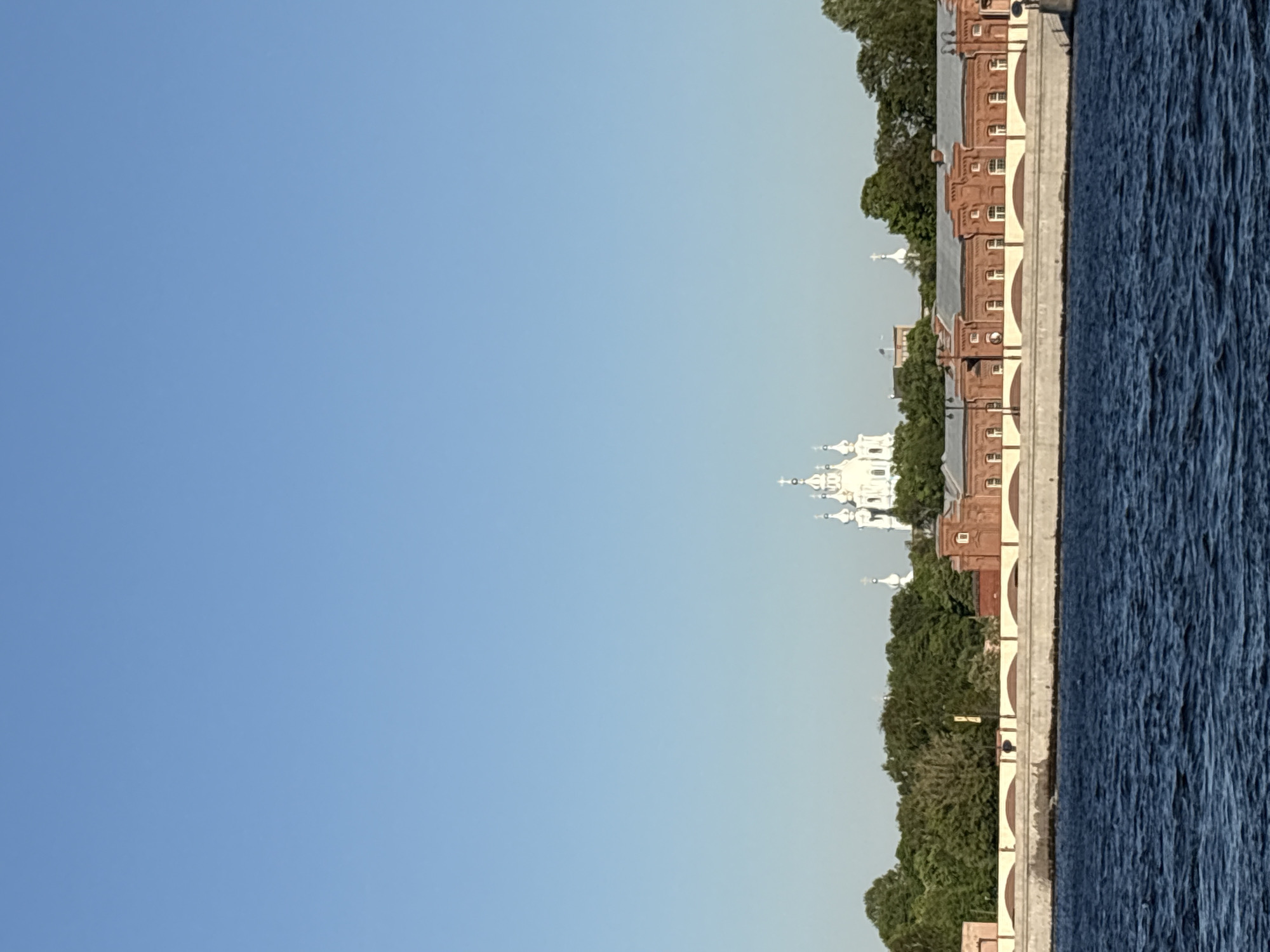 The image depicts a waterfront view in Saint Petersburg, Russia. A body of water, likely the Neva River, is on the right side of the image. Along the embankment are red brick buildings, interspersed with trees, suggesting a historical or governmental district. A white church or cathedral with multiple domes is visible among the buildings, characteristic of Russian Orthodox architecture. The sky is clear and blue, occupying the left side of the frame. The scene suggests a calm, sunny day in a historic part of Saint Petersburg.