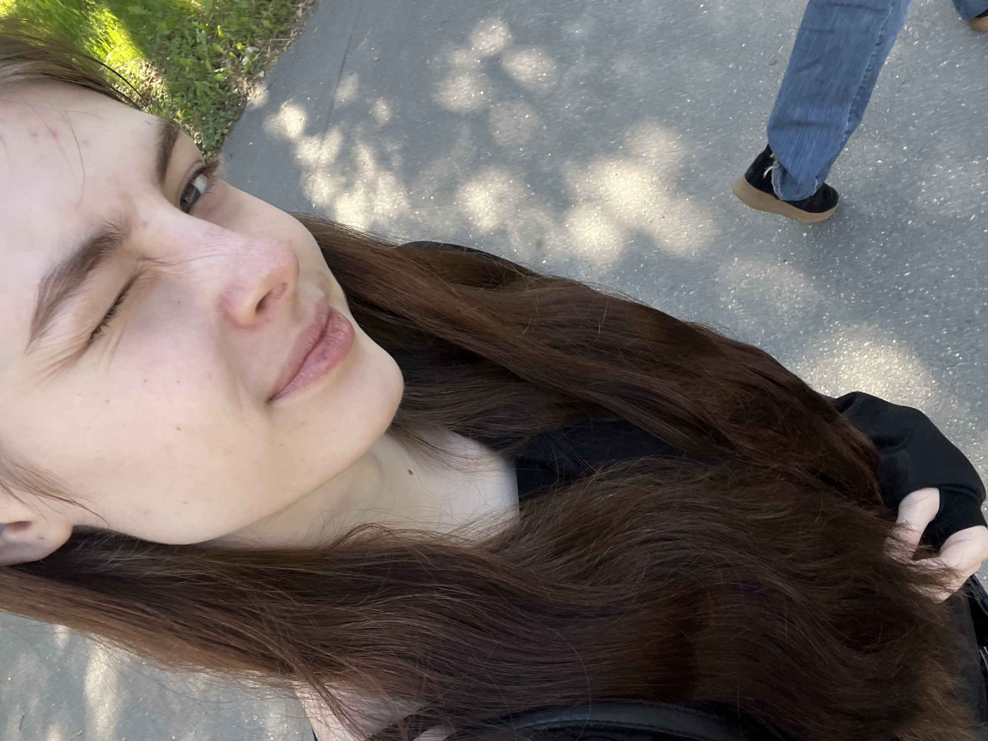 The image captures a playful selfie taken outdoors by a young person, who appears to be female. She is positioned diagonally in the frame, with her long, dark brown hair cascading around her shoulders. She winks with her right eye closed and has a slight smile on her lips, conveying a cheerful and lighthearted mood. Her left eye is open, looking upwards towards the camera. She wears a dark-colored top or jacket.
The background shows a paved surface, likely a sidewalk or path, with dappled sunlight and shadows from overhead foliage, suggesting a sunny daytime setting. To the right, the lower leg and foot of another person are visible, indicating their presence nearby. This person is wearing blue jeans and black shoes with light brown soles, appearing to be in motion. On the far left side of the frame, vibrant green grass and some natural foliage are visible, suggesting a park or green space. There is no visible text in the image.