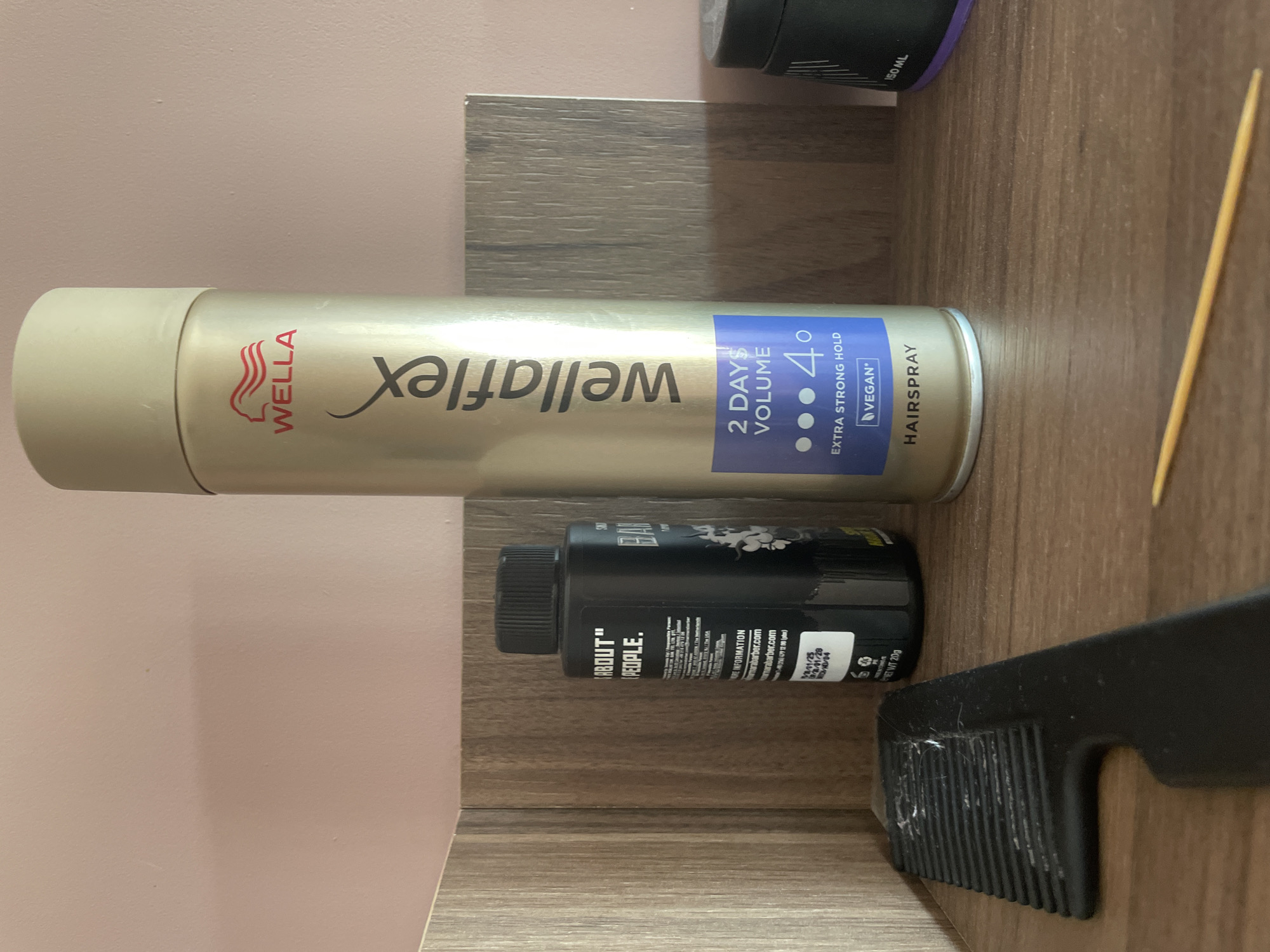 The image displays a collection of personal care and miscellaneous items resting on a wooden surface, with a light pink or beige wall in the background, likely in Ihtiman, Bulgaria.
The most prominent object is a silver Wella Wellaflex hairspray can, positioned horizontally across the center. Its label features the "WELLA" logo in red, the "wellaflex" brand name, and a blue section indicating "2 DAYS VOLUME," "4° EXTRA STRONG HOLD," and "VEGAN HAIRSPRAY."
Below the hairspray can, a black bottle, possibly hair styling product, is also lying horizontally. Visible text on this bottle includes "ABOUT" "PEOPLE.", the brand name "BAR", "MORE INFORMATION," "barabarber.com," and "NET WT 200." Date codes such as "M01/25," "01/28," and "D/04" are also present.
In the bottom right corner, a black comb with wide teeth is partially visible, lying on the wooden surface. It appears to have some white lint or hair caught in its teeth. A thin, light-colored wooden stick, resembling a toothpick or skewer, is positioned diagonally in the mid-right area.
At the top right, a small portion of a dark bottle, possibly another personal care item, is visible, showing the text "150 ML." The wooden surface exhibits a noticeable grain pattern, suggesting a shelf or countertop in an indoor setting. No specific activity is taking place; the image is a still life of these everyday objects.
