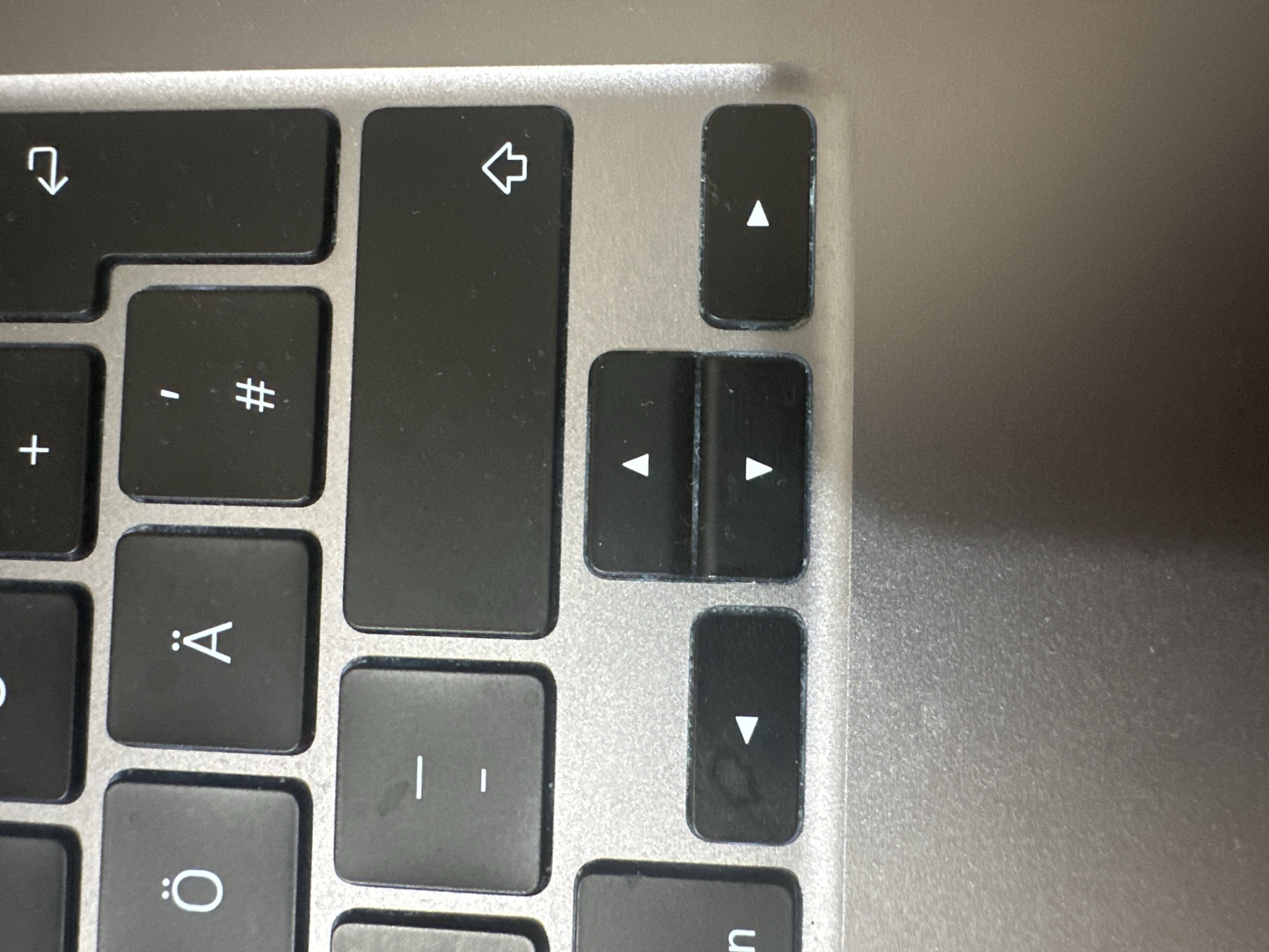 The image is a close-up, high-angle shot of a portion of a laptop keyboard. The keyboard is dark gray with white lettering. Visible keys include those with the characters "Ö", "Ä", a dash "-", and an upward-pointing arrow. There are also arrow keys: left, up, down, and right. The laptop casing appears to be metallic silver. The background is blurred, but suggests a smooth, possibly slightly textured surface. There are no people, actions, or interactions visible. The scene is an indoor, desk-like environment, with no discernible connection to the city of Hamburg, Germany.
