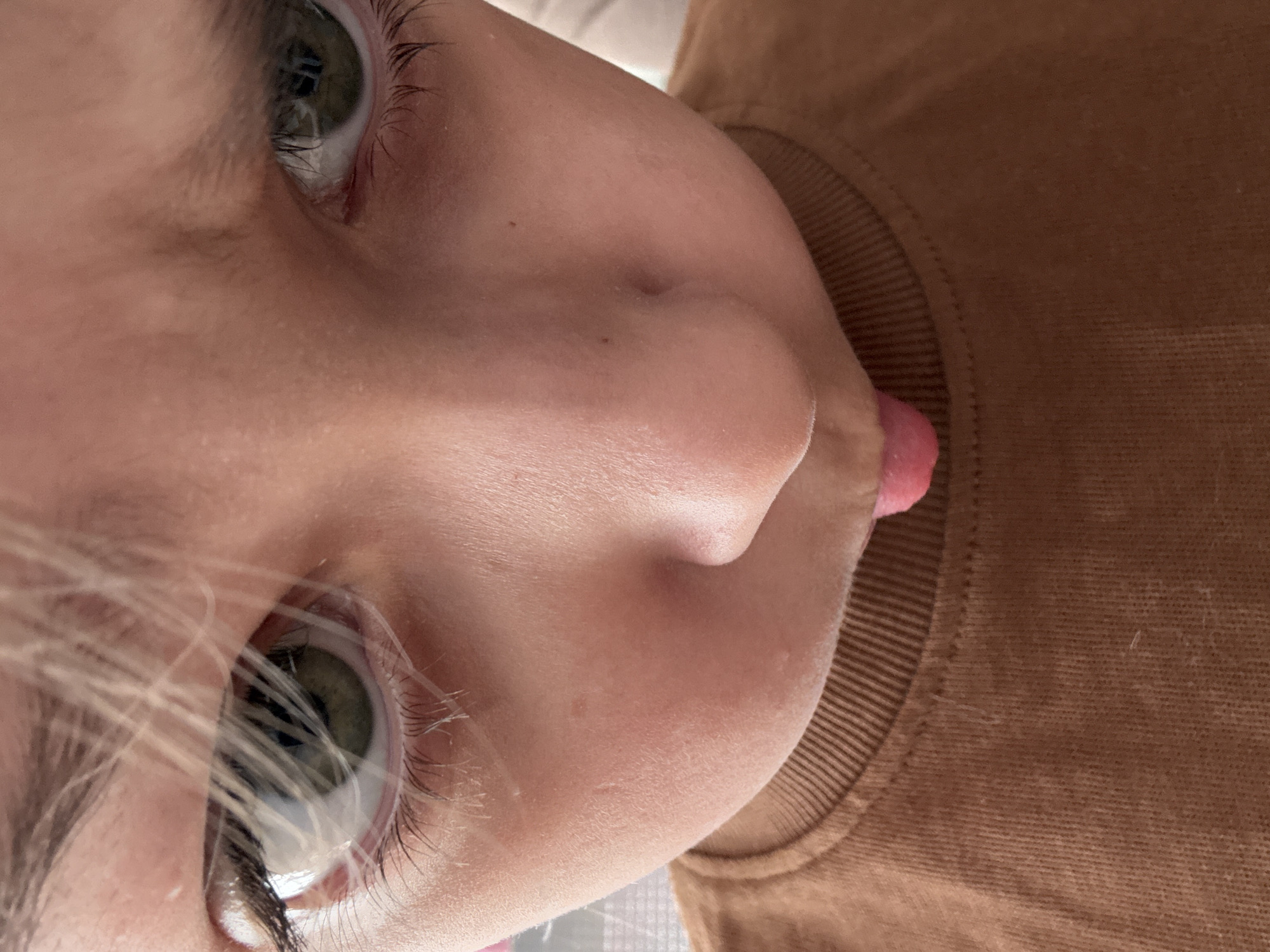 A close-up, tilted shot depicts a child's face and part of their upper body. The child has fair skin, blonde hair, and green eyes with visible eyelashes. Their nose is prominent in the frame, and a pink tongue is slightly visible between their lips. The child is wearing a brown, ribbed-neck sweater or shirt. The background is blurry and indistinct. There's no information provided in the image that would suggest a connection to Votkinsk, Russia.