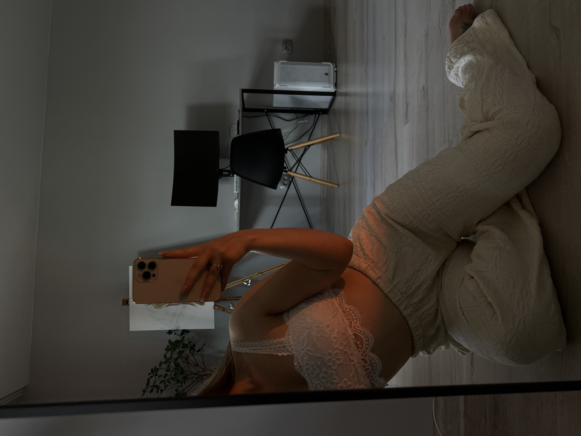 A person is taking a selfie in a mirror. They are wearing a white lace bralette and cream-colored patterned pants. The person is sitting on the floor, with their legs crossed. Their right arm is extended, holding a gold smartphone to take the photo. A ring is visible on their right hand.
The background shows a modern room with a light gray wall and a wooden floor. In the reflection, there is a black desk with two computer monitors, a black chair with wooden legs, and a white rectangular object on a shelf. A small plant with green leaves is visible in the lower left corner of the mirror's reflection. The lighting suggests it might be daytime, with natural light entering the room. There is no visible text in the image. The setting appears to be an indoor domestic space, possibly a home office or living area. The overall mood is casual and relaxed. The location context is Ternopil, Ukraine.