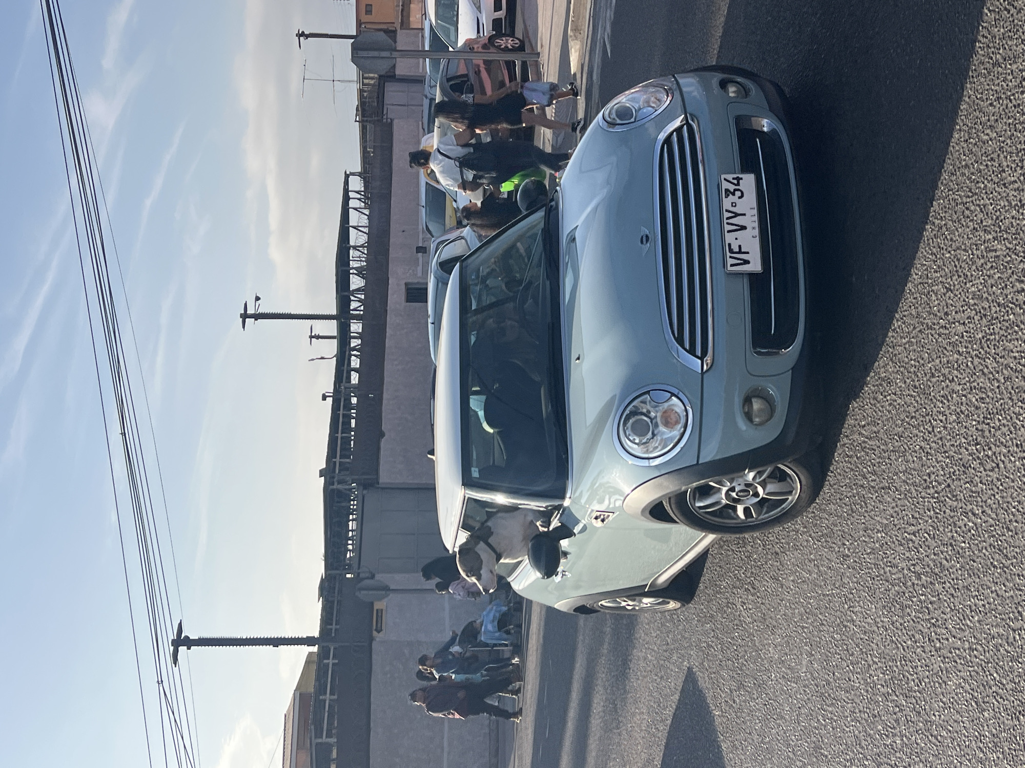 In Arica, Chile, a light blue Mini Cooper is captured in motion on a paved road. A dog with tan markings is looking out the driver's side window, its head and shoulders visible. Behind the Mini Cooper, several people are walking along a sidewalk next to a grey concrete wall and a metal fence with a covered walkway. The sky is clear blue with some wispy clouds, and utility poles with wires are visible in the background. The license plate of the Mini Cooper reads "VF-VY-34 CHILE".