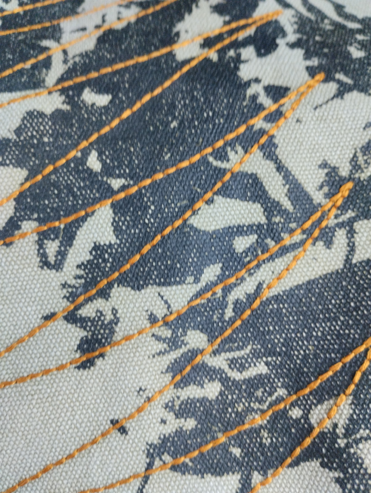 The image is a close-up shot of a textured fabric with a dark gray and white pattern, overlaid with thin, parallel orange stitches. The fabric appears to be a canvas or a similar coarse material, with visible woven threads creating a subtle grid. The pattern on the fabric is abstract, resembling splashes or organic shapes in dark gray against a lighter background. The orange stitches run diagonally across the frame from the bottom left to the top right, creating a series of equidistant lines. The lighting is even, and the focus is sharp on the fabric's texture and the stitching. There are no people, animals, or identifiable landmarks visible in the image. The image does not provide any information that would allow for its location to be specified within Castione Andevenno, Italy, or any other geographical area.