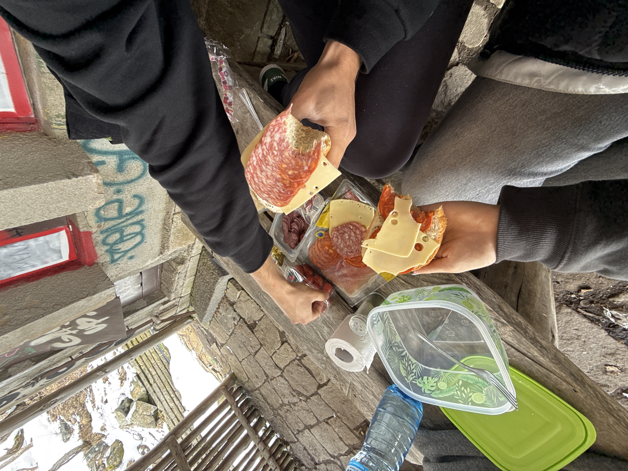 Captured from an elevated perspective, the image shows a picnic scene featuring three people sharing a meal in what appears to be an outdoor setting. The picnic is arranged on a weathered wooden surface, possibly a bench or table, and includes a spread of bread, sliced salami, and cheese. One person is assembling a sandwich with these ingredients.
Additional items on the wooden surface include a package of salami and cheese, a roll of toilet paper, a water bottle, and a clear plastic container with a green lid and a fork inside.
The setting is a stone structure that looks like a small building or a lookout point. There is graffiti on the stone walls. Through an opening, one can see a snowy landscape below.
The location of this picnic is near Sofia, Bulgaria, based on the appearance of the landscape and the architectural style. It might be near or in Vitosha mountain.