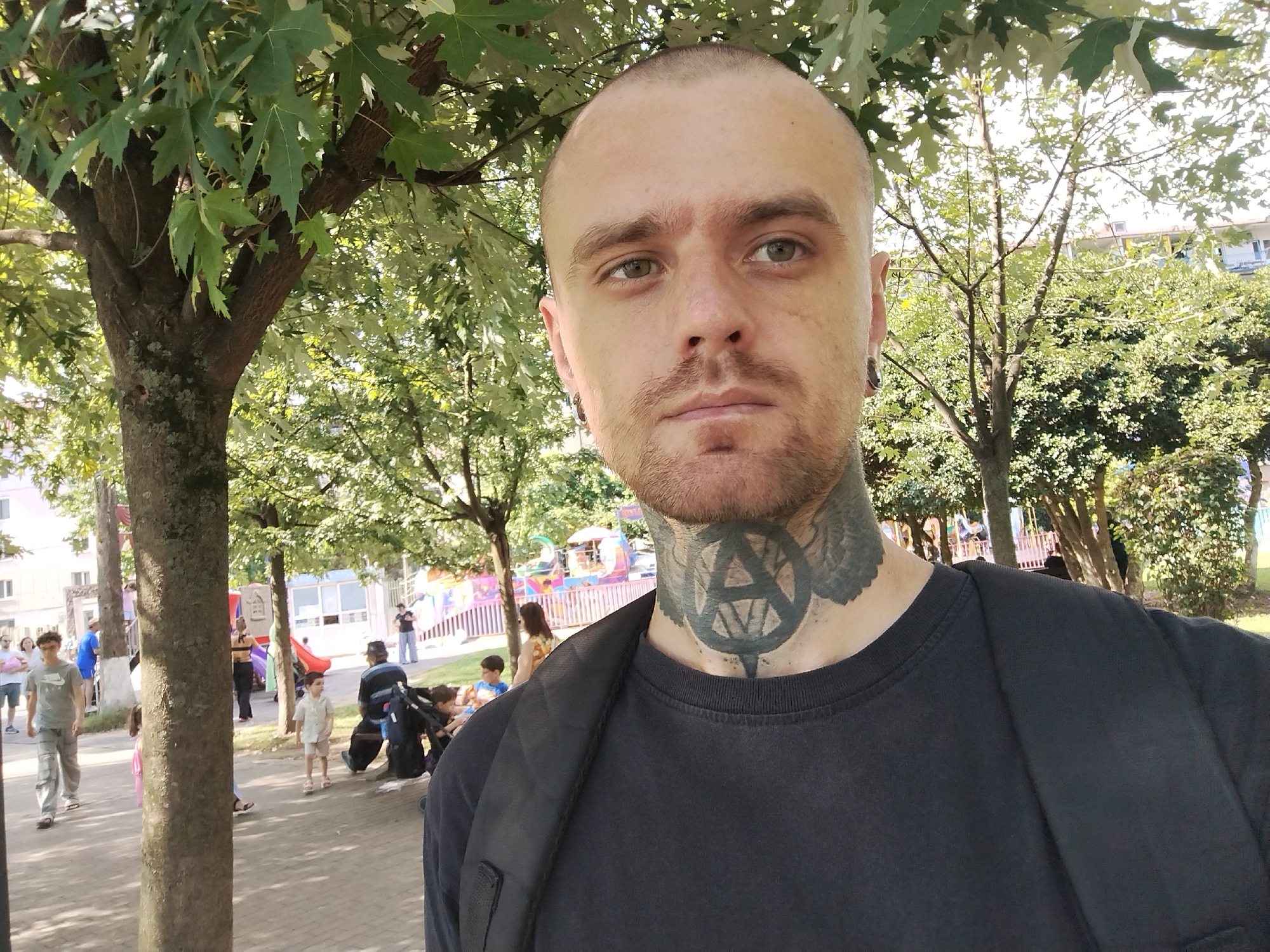 A selfie taken outdoors features a bald man with a shaved head, a short beard, and a tattoo on his neck. He is wearing a black t-shirt and a black backpack. Behind him, trees with green leaves provide shade. In the background, several people are visible, including children and adults, in what appears to be a park or public area with playground equipment and buildings in the distance. The overall scene suggests a sunny day in a casual urban setting.