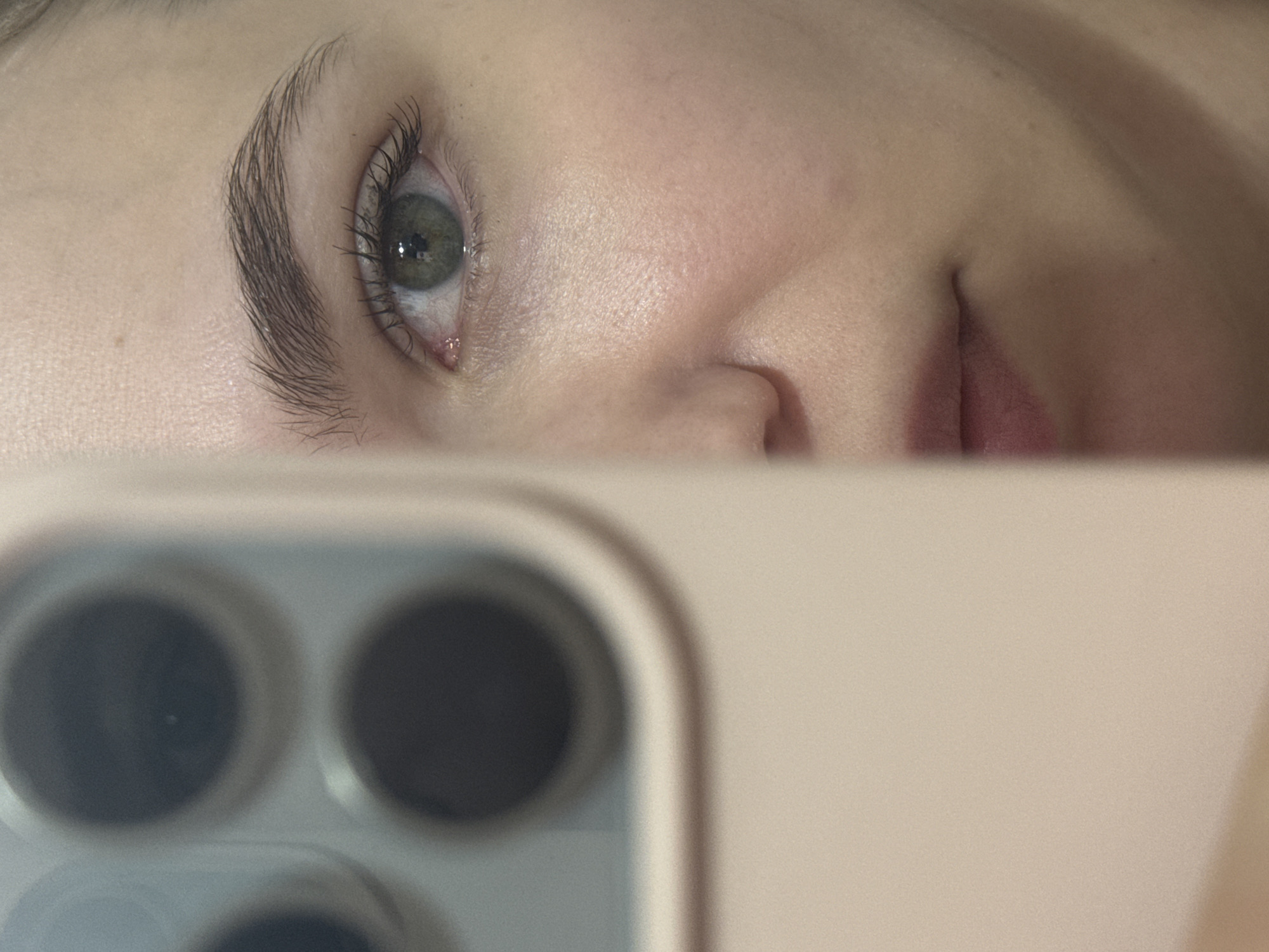 This close-up, diagonal shot captures the upper right side of a person's face, primarily focusing on a single eye, partially obscured by a smartphone in the foreground. The person's eye is open, revealing a vibrant green iris with subtle hints of hazel and a dark pupil reflecting a light source. Eyelashes are visible, appearing long and dark, possibly enhanced with mascara, and the eyebrow is well-defined. The skin on the cheek and around the eye is fair, showing natural texture and pores, with a slight redness near the tear duct. Part of the nose is visible towards the right side of the face. In the blurred foreground, a light pink or beige smartphone case is prominent, showing the out-of-focus circular lenses of its multi-camera system. The overall composition suggests the person is looking into the phone, possibly taking a selfie or engaging with the screen. The setting is likely indoors in Chaikovsky, Russia, with soft, even lighting. No text is visible.