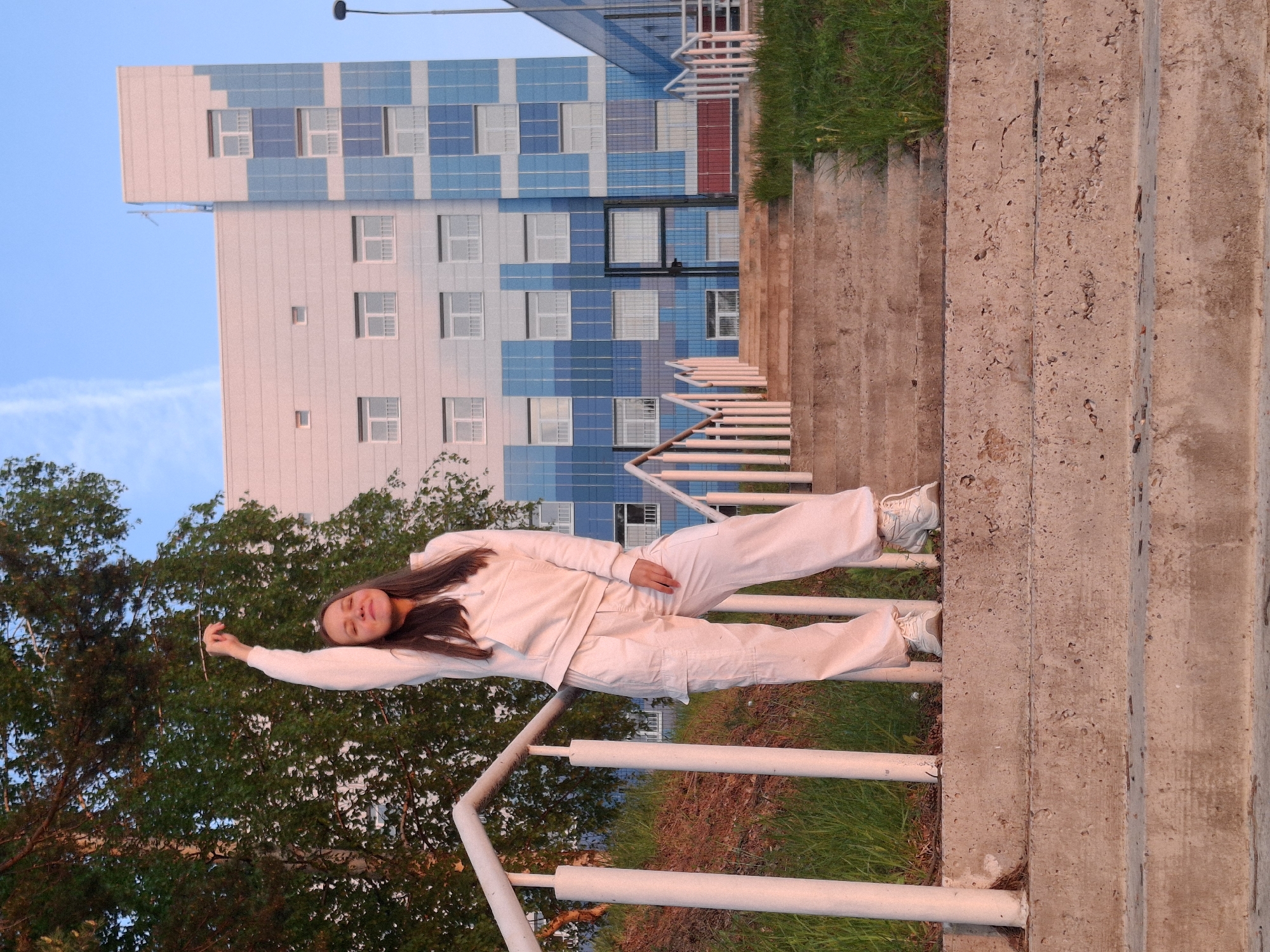 A young woman in white attire is reclining on the handrail of a staircase in Chaikovsky, Russia. She is facing the camera with her left arm extended upward. In the background, a modern building with a blue and white facade is visible, along with greenery, including trees and grass. The setting is likely an urban park or public space.