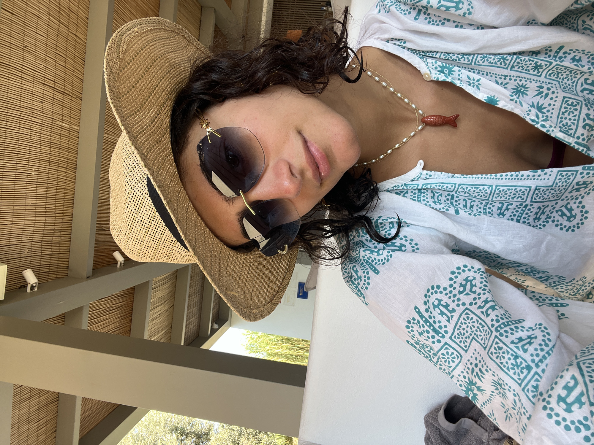 A young woman with dark curly hair wears a straw hat and oversized sunglasses. She is dressed in a white and teal patterned tunic and a pearl necklace with a red fish pendant. The background shows a covered outdoor area with a bamboo ceiling and supporting pillars. The scene appears to be a casual setting, possibly a resort or a waterfront cafe in Naousa, Greece.