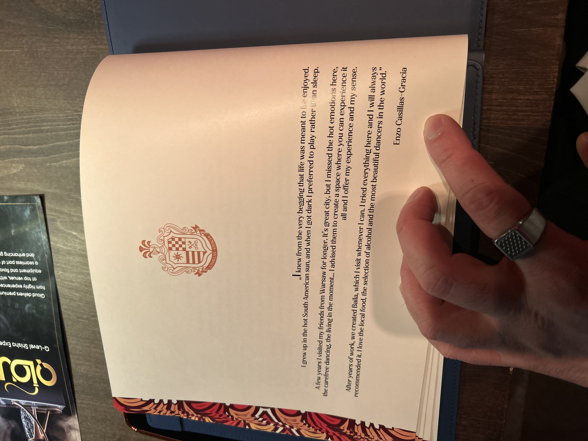 The image shows an open book with text and a coat of arms-like crest on the pages. The text seems to be a quote by Enzo Casillas-Gracia, mentioning his enjoyment of life, growing up in the South American sun, and a connection to Warsaw. He mentions missing the hot emotions and carefree dancing in Warsaw, inspiring him and his friends to create "Baila" - a space to experience these things. He also professes his love for the local food, alcohol, and the most beautiful dancers in the world at Baila. The location of Baila is in Warsaw, Poland. A hand with a ring is visible on the right side of the book. Part of a brochure titled "Qloud-Level Shisha Experience" is also visible next to the book. The environment appears to be a table or a similar flat surface.