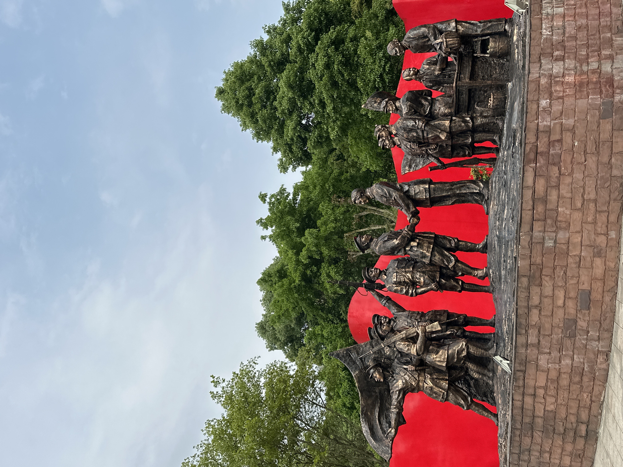 The image shows a bronze-colored monument featuring a group of figures set against a bright red backdrop and a brick-like base. The figures appear to be soldiers or revolutionaries, depicted in various poses with weapons and tools. A large tree is visible behind the monument, and the sky is overcast. The monument's location is in the Liangjiang New Area of Chongqing, China.
