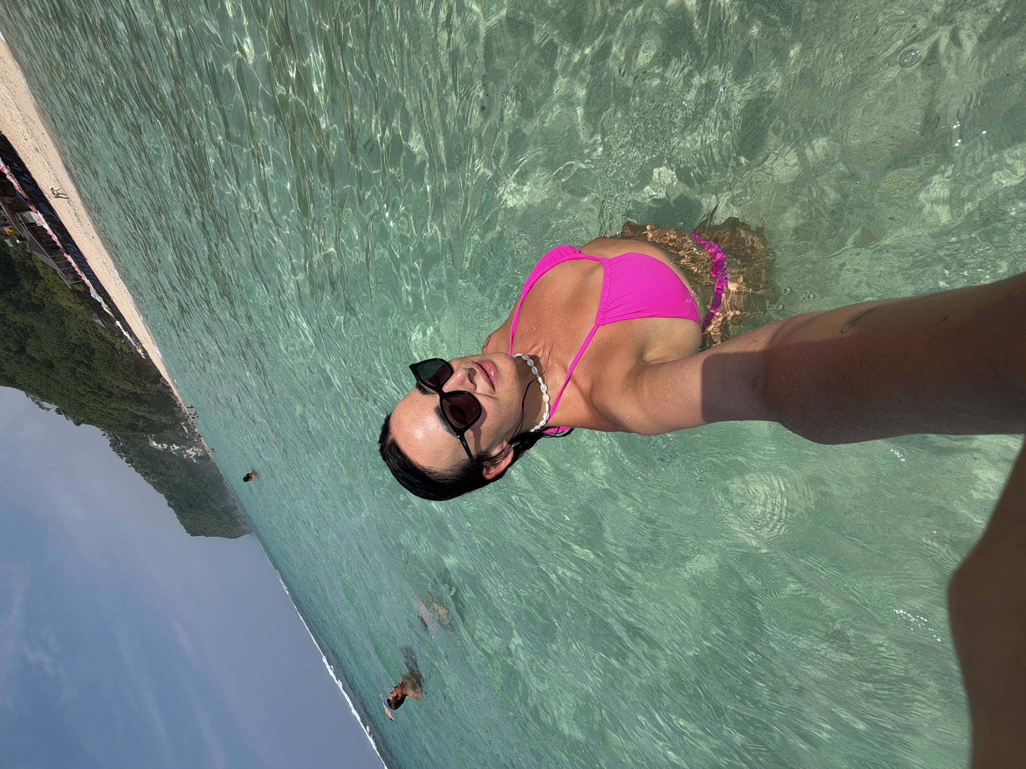 A female subject with dark, wet hair, sunglasses, and a white shell choker necklace is captured in a selfie-style photograph. She wears a bright pink bikini top and matching bottoms, and is partially submerged in clear, shallow, turquoise ocean water, which reaches approximately chest height. Her right arm, showing a faint tattoo near the elbow, extends forward, holding the camera.
In the background, to the left of the main subject, a male individual is partially submerged in the water, looking towards the right. Multiple other indistinct figures are visible further in the water and on the sandy beach. The beach extends to the right, backed by a large, steep, green-vegetated cliff face. Along the base of the cliff, on the beach, are several structures resembling beach shacks with colorful awnings (red, blue, striped). The sky is a clear blue with minimal cloud cover. The scene depicts a sunny day at a beach in Ungasan, Indonesia.
