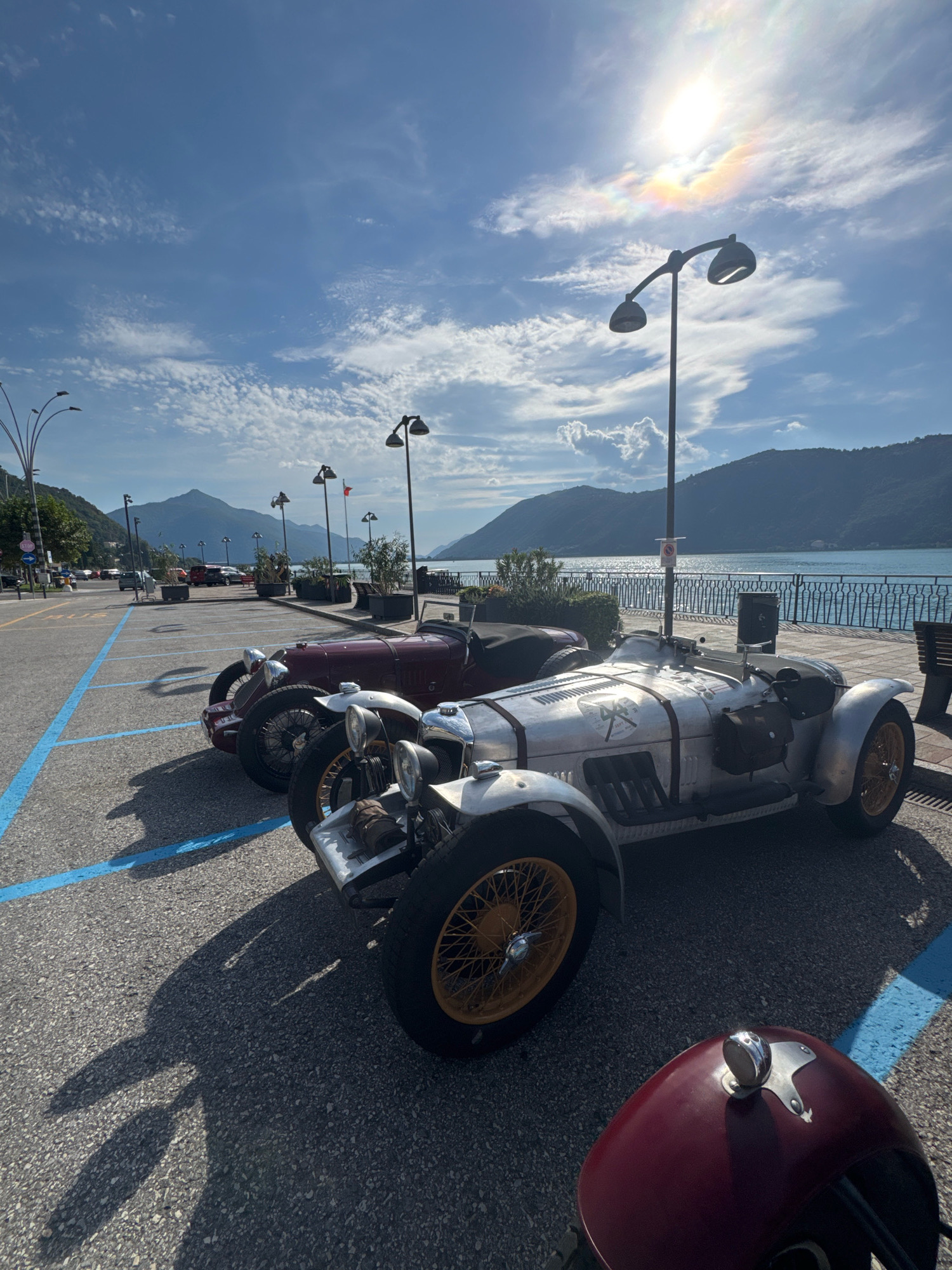 The image captures a sunlit scene in Campione d'Italia, Italy, along the shore of a lake. In the foreground, three vintage cars are parked on asphalt marked with blue parking lines. The car closest to the viewer is partially visible, showing a burgundy fender with a chrome embellishment. Behind it, a silver vintage car with yellow spoked wheels is prominently displayed, featuring a streamlined body, leather straps, and side-mounted storage bags. To the left of the silver car is another vintage car, this one a deep burgundy color, with its hood open and wire-spoke wheels visible.
The background reveals a body of water, likely Lake Lugano, reflecting the bright sunlight. A promenade lined with a metal railing and benches runs along the waterfront, with several lampposts providing illumination. Beyond the lake, rolling green hills and mountains form a picturesque backdrop under a clear blue sky dotted with wispy white clouds. The sun is high in the sky, creating a strong glare and a faint rainbow effect. The overall atmosphere suggests a pleasant day, possibly during a classic car event.