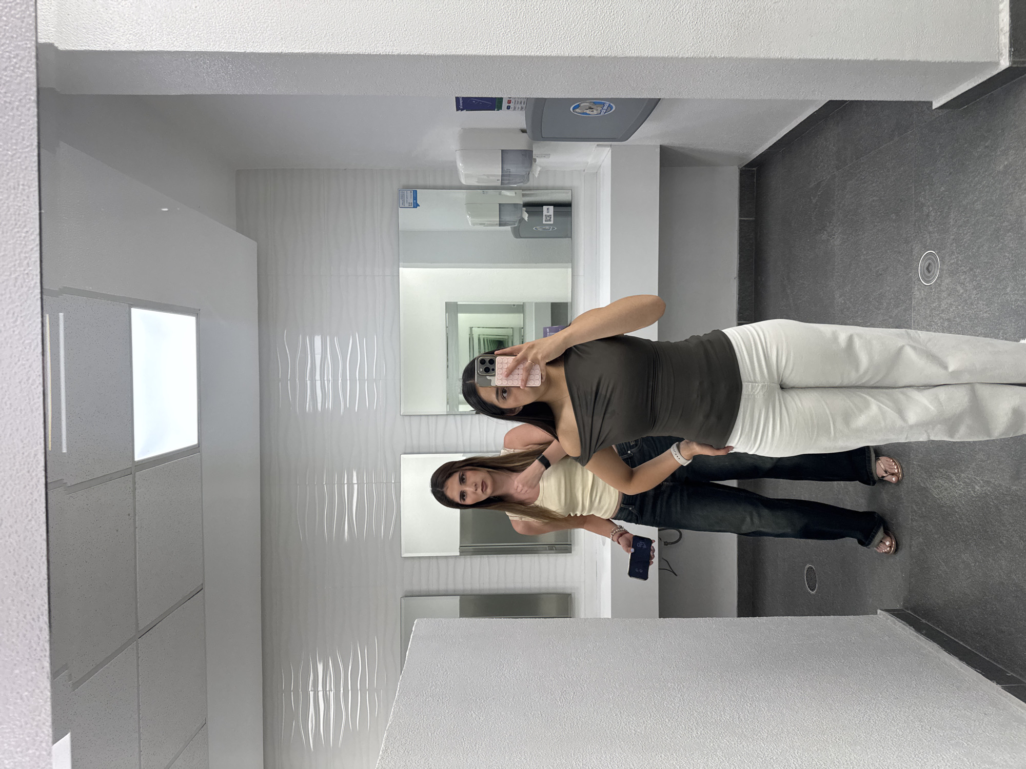 This media file captures a mirror selfie of two young women in a modern, well-lit public restroom. The image, originally rotated 90 degrees clockwise, shows the women standing in front of a counter with mirrors above it.
The woman taking the selfie has dark hair and is wearing a dark olive green top and white pants. She holds a smartphone with a pink bubble-textured case, reflecting her face and upper body. Her right hand is raised, possibly making a "V" sign, though it's partially obscured by the phone.
Standing next to her, also reflected in the mirror, is another woman with light brown hair. She wears a light yellow or cream-colored tank top and dark jeans. She is giving a thumbs-up gesture with her right hand and holds a dark smartphone in her left hand, which displays "6:20" and "67%". Both women appear casual and cheerful, posing for the photo.
The restroom features clean, contemporary design elements. The walls behind the women are adorned with white, wavy-textured tiles. Above the counter, on the back wall, are a hand soap dispenser, a hand dryer, and a baby changing station. The baby changing station has a blue sticker with a stylized baby logo, likely from "KOALA KARE". The floor is composed of dark gray tiles, and the ceiling features white acoustic panels with integrated rectangular light fixtures. The overall setting is brightly lit by overhead fluorescent-style lighting. This location is situated in Puebla, Mexico.
