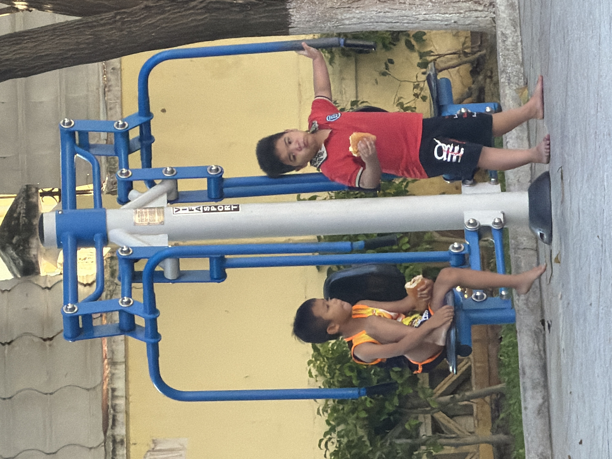 The image shows two young boys standing near an outdoor exercise machine in a public area. The boy on the left, wearing an orange and yellow sleeveless shirt and black shorts with orange trim, is seated on the exercise machine, holding a piece of food in his hands. The boy on the right, wearing a red polo shirt and black shorts with white lettering, is standing and also holding a piece of food. They appear to be in a park or recreational area, with a yellow wall in the background and a tree to the right. The exercise machine is blue and gray. The ground is paved. The scene suggests a casual outdoor setting where children are present.