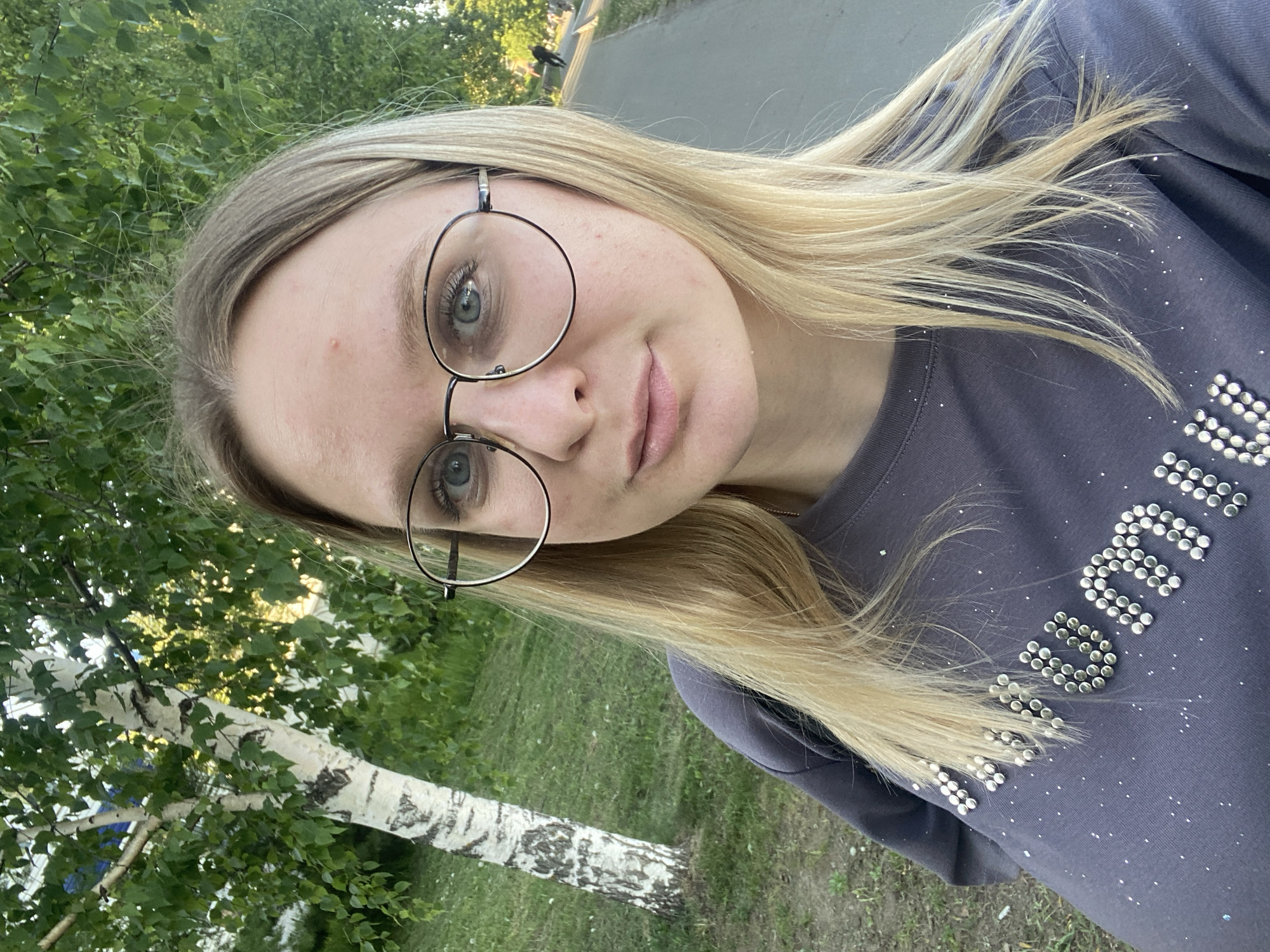 A blonde woman with glasses stands outdoors. The background features a birch tree and green grass. The woman is wearing a gray shirt with silver studs. The location is Unknown, Unknown.