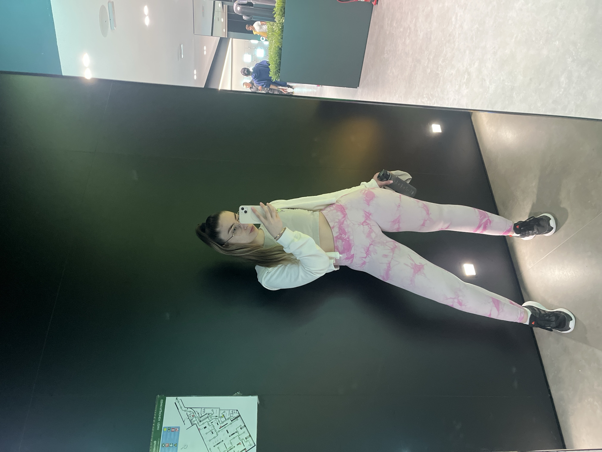 The image shows a woman taking a selfie in what appears to be a modern, well-lit interior space. The woman is wearing a light-colored top, a white hoodie, and pink tie-dye leggings with black and white sneakers. She's holding a phone in her right hand, presumably to take the picture. She is also holding a black water bottle. Behind her is a dark wall with a building map posted on it. In the background, other individuals can be seen further back in the space. It is unknown where this picture was taken.