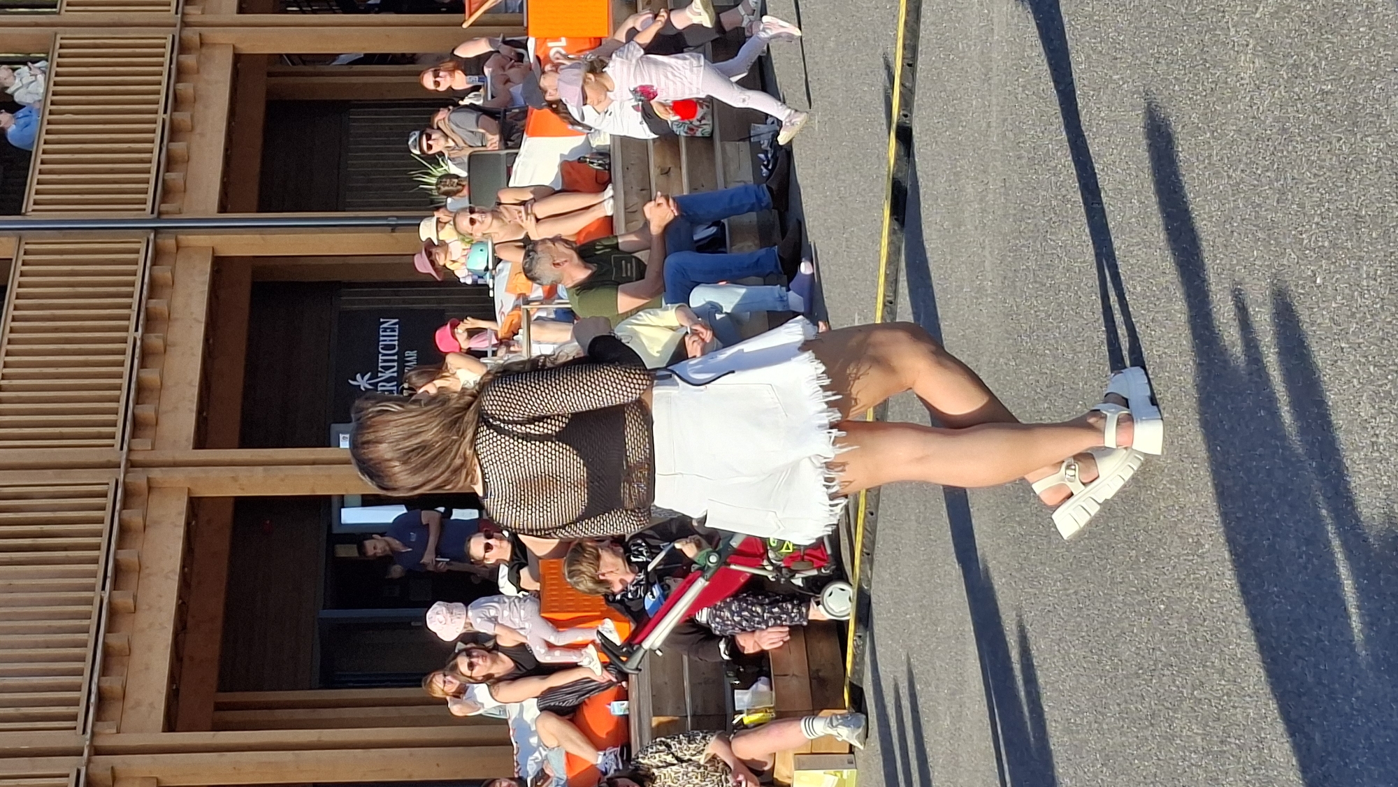 The image depicts an outdoor scene, possibly a market or public gathering area, with a wooden structure in the background that appears to be part of a building with multiple levels and railings. Several individuals are visible, some seated on tiered wooden seating and others standing. In the foreground, a woman is seen from the back, wearing a black fishnet top over a black bra, and white denim shorts with a frayed hem. She is also wearing white platform sandals. To her right, a young child in a pink striped outfit and a pink hat is running. The ground is paved with asphalt, and shadows from the overhead structure and people are cast upon it. The setting appears to be a sunny day. While the location is stated as Tallinn, Estonia, no specific landmarks or identifying features within the image confirm this.