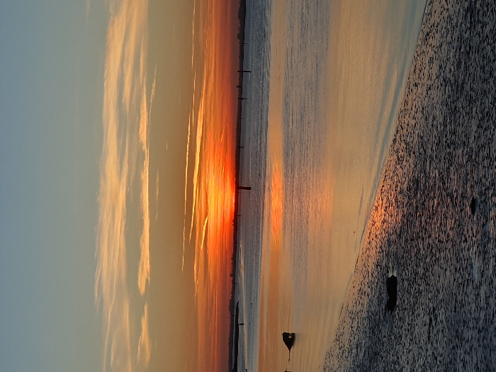 The image displays a panoramic view of a waterway, identified as Breydon Water or the Great Yarmouth Estuary in Great Yarmouth, United Kingdom. The scene is set at either sunrise or sunset. A vibrant orange-red sun is partially visible on the distant horizon, illuminating the sky with intense orange, red, and yellow tones that gradient into a light blue upper atmosphere. Thin, horizontal clouds are present across the sky. The water surface is calm, exhibiting a prominent, elongated reflection of the sun's light stretching towards the foreground. The immediate foreground features a textured, wet shoreline, likely composed of sand or mudflats, diagonally extending from the lower left. Two dark, undefined objects, consistent with debris, are situated on this shoreline. Numerous dark, slender, vertical posts or buoys are interspersed across the water, suggesting a marked channel. One distinct reddish buoy is visible near the sun's reflection. No individuals are depicted in the image.