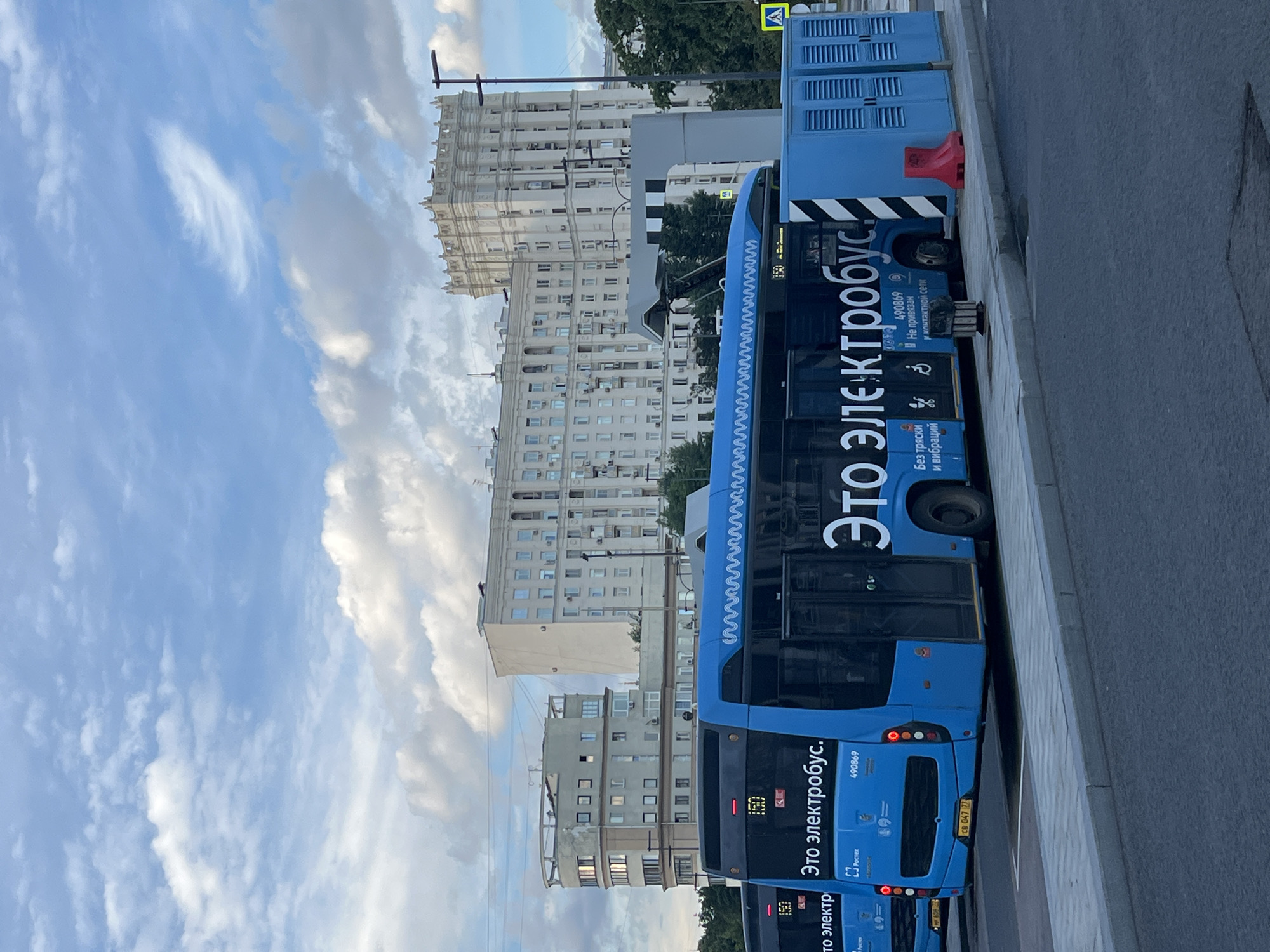 This is a vertical, eye-level shot of a blue electric bus parked on a city street. The bus is the primary subject, with its large, bold Cyrillic text reading "Это электробус" (This is an electric bus) prominently displayed along its side. The text also mentions "Без тряски и вибрации" (Without shaking and vibration), suggesting a smooth ride. Other text on the bus includes the number "490869" and the logo "Ростех".
Behind the bus, a row of tall, pale buildings with numerous windows and balconies dominates the background. These buildings appear to be apartment blocks, characteristic of urban architecture. The sky above is partly cloudy with streaks of white clouds against a light blue backdrop, indicating daytime, possibly late afternoon or early evening given the lighting.
The bus itself has its doors closed, and a red traffic barrier is visible next to it. The street in the foreground is paved, with a sidewalk and a curb. The overall scene suggests an urban environment in Moscow, Russia, as indicated by the Cyrillic text and architectural style. The atmosphere is calm, with no visible people, and the focus is on the modern public transportation vehicle and its surroundings. A license plate is partially visible, showing "CB 047".