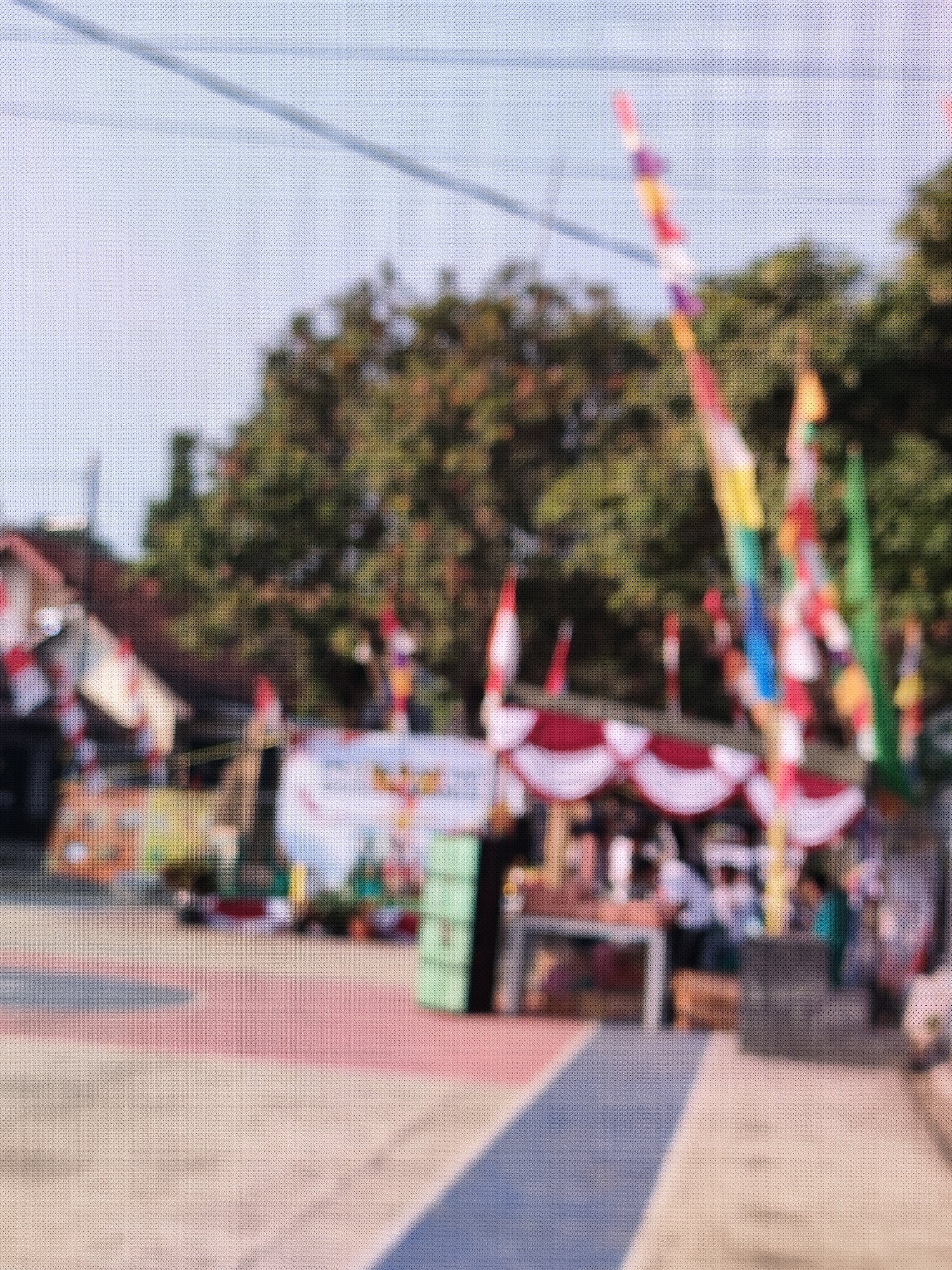 The image depicts an outdoor scene with several elements suggesting a public gathering or event. In the background, trees and buildings are visible. Colorful flags, some with red and white patterns, are prominently displayed, indicating a festive or celebratory atmosphere. There are also what appear to be market stalls or booths set up, with people gathered around them. The ground in the foreground has a patterned surface, possibly a paved area with colored lines. The overall image is blurred, suggesting it was taken from a distance or with a shallow depth of field. It is impossible to determine the location in the city of Caracas, Indonesia, as Caracas is the capital of Venezuela, not a city in Indonesia.