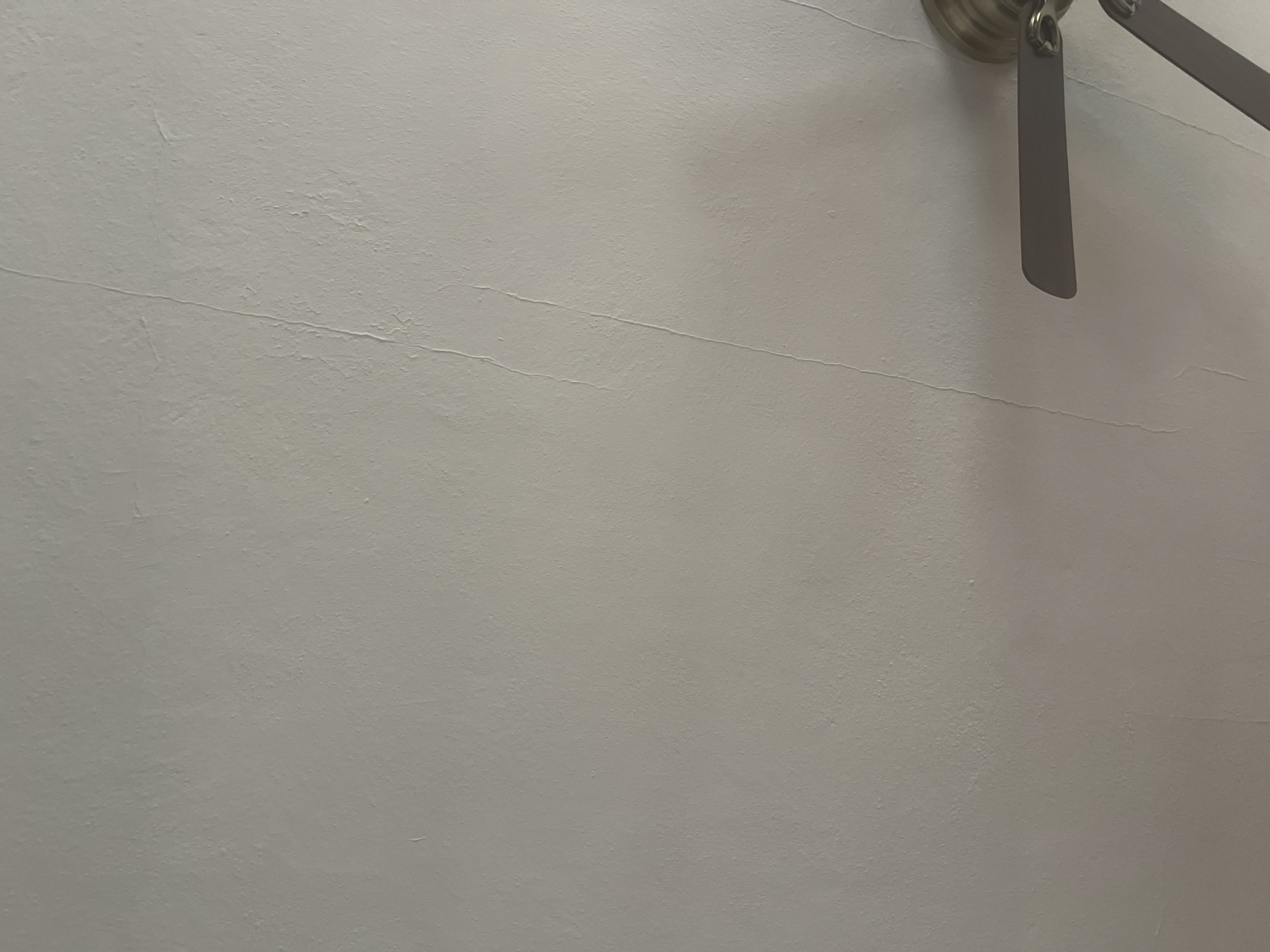 The image shows a close-up view of a textured white ceiling with a faint crack running vertically down the center. A portion of a ceiling fan, with brass-colored housing and dark blades, is visible in the lower right corner. The scene appears to be an indoor residential setting, likely a bedroom or living room. The lighting is dim, casting a shadow from the fan onto the ceiling. No people are present in the image. The location is identified as West Hartford, United States of America.