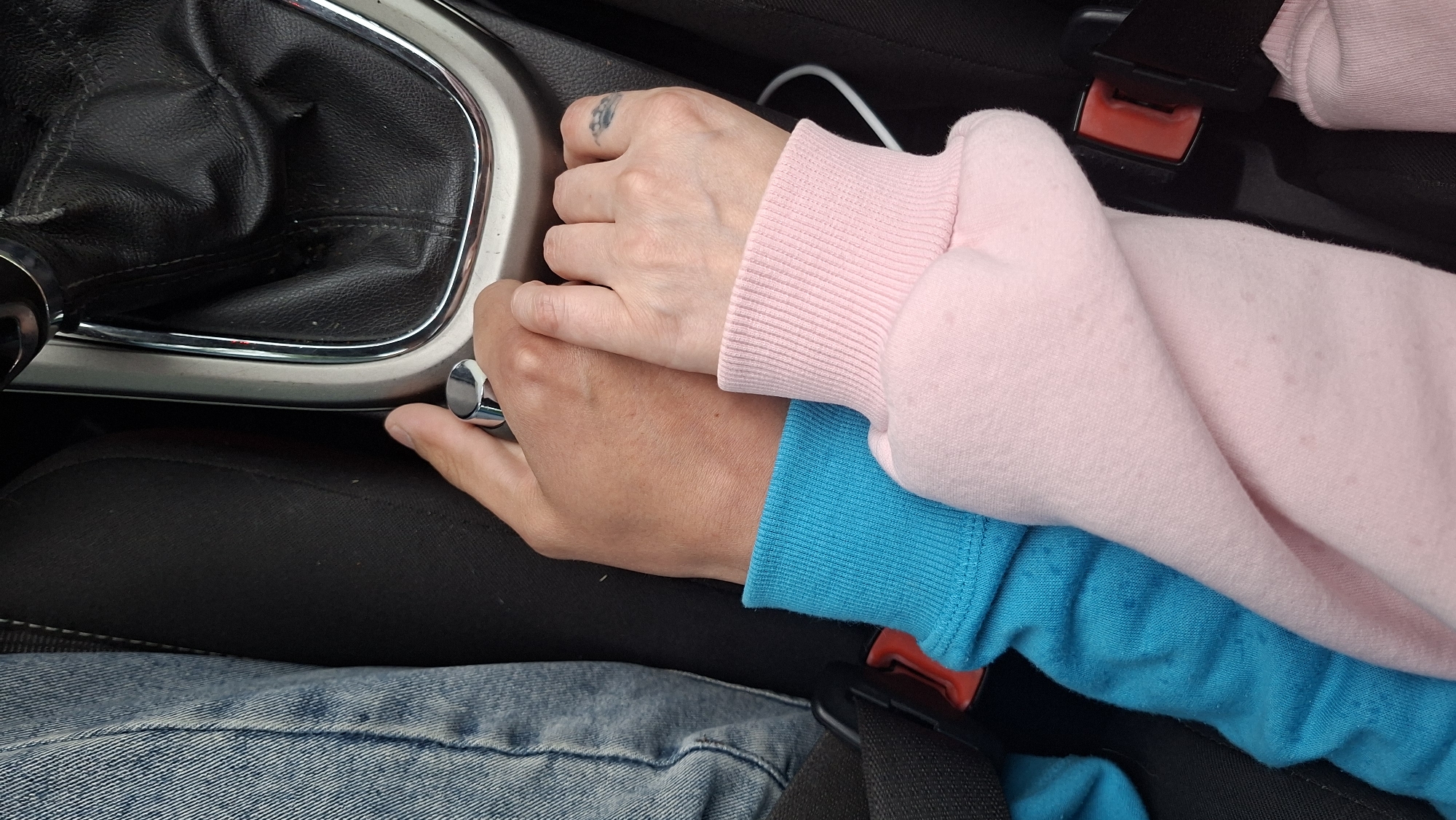 Two people are holding hands in the front passenger seat of a car. The person on the left is wearing a blue sweatshirt, and the person on the right is wearing a pink sweatshirt. Their hands are clasped, and their arms are wrapped around each other. The car's gear shifter is visible in the background. The image appears to have been taken from a low angle, looking up at the hands and arms. The scene is well-lit, with natural light coming from the right side of the frame.