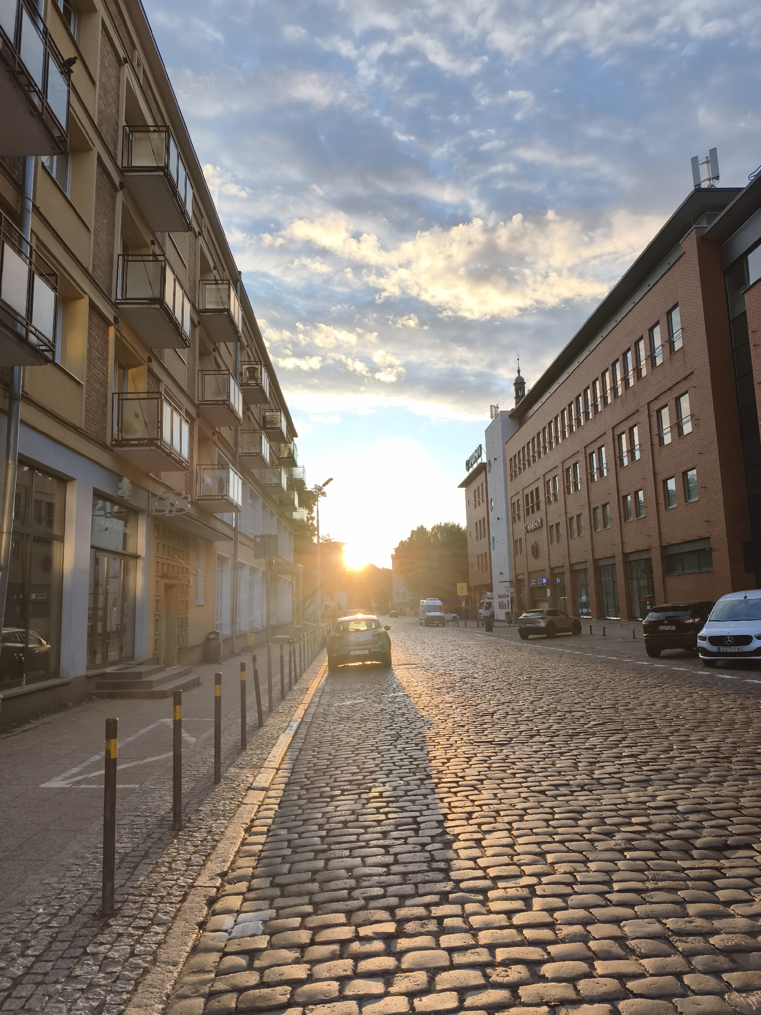 The image depicts a street scene in Gdańsk, Poland, during sunset. The sun is low on the horizon, casting long shadows and illuminating the cobblestone street and the facades of the buildings. On the left, a multi-story building with balconies lines the street. On the right, a brick building with numerous windows stands. Several cars are parked or driving on the street. The sky is filled with clouds, some of which are lit by the setting sun. The overall impression is that of a European city street bathed in the warm glow of the evening sun.