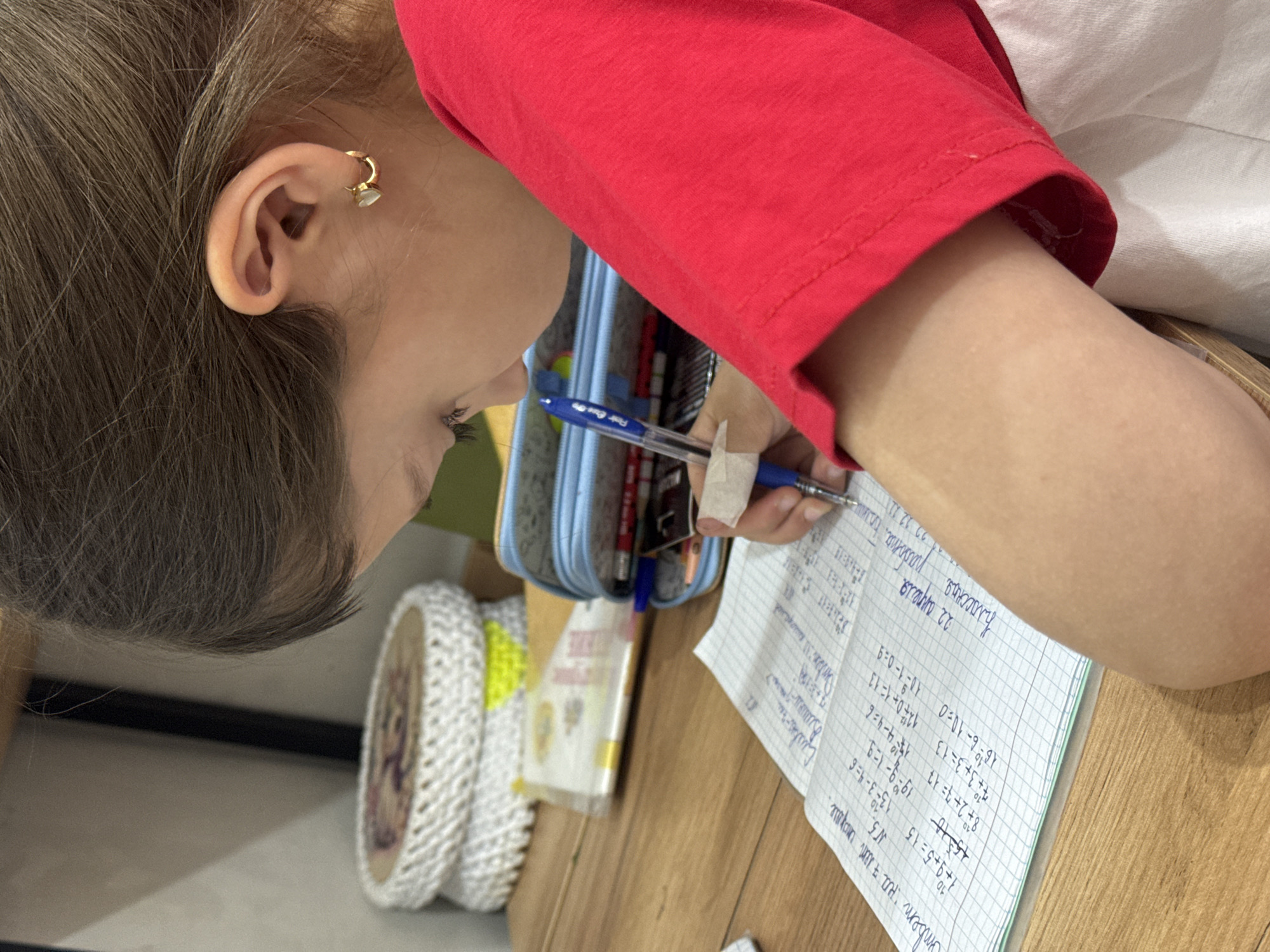 An overhead close-up shot captures a person, likely a child or young adult, with light brown hair and a red short-sleeved top, focused on writing in a notebook. The scene is indoors, with the person seated at a wooden table.
The main subject is seen from a high angle, looking down at the notebook. A small, gold-colored hoop earring with a white element is visible on their right ear. Their arm, wearing a red shirt, extends across the frame as they hold a blue ballpoint pen, which has white tape wrapped around its grip, to write on a lined notebook page. A light blue pencil case is partially visible behind the pen.
The activity clearly depicts someone engaged in schoolwork or homework. The notebook features graph paper and is filled with handwritten Russian text and mathematical equations. Visible text includes "22 апреля" (April 22), "Классная" (Classwork), and various arithmetic problems such as "1+9+5=15", "8+2+7=17", "7+3+3=13", "12+0+1=13", and "10-1-0=9". Some numbers appear crossed out. A phrase "ответ на 7 лет сноуде" (answer to 7 years of Snowden) is also present. The pen itself has text "Fair Excep" visible, possibly part of a brand name.
The setting appears to be a study area or desk, with other blurred objects, including a white, woven-textured item, visible in the background. The use of Russian text aligns with the provided location context of Votkinsk, Russia. The time of day and weather are not discernible. The person's expression is one of concentration.