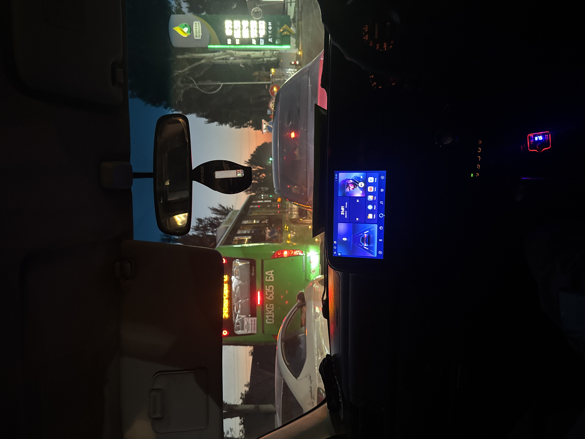 The image shows a view from inside a car in Bishkek City, Kyrgyzstan, at dusk. Traffic is heavy, with a green bus in front displaying the license plate "01KG 605 BA" and "243 бишкек". A white car is to the left of the bus. The car interior is dark, with a glowing infotainment screen displaying a music player interface. A red-lit device is visible below the screen. In the background, a gas station with a "BISHKEK PETROLEUM" sign is illuminated, with prices displayed in local currency. The sky is tinged with sunset colors.