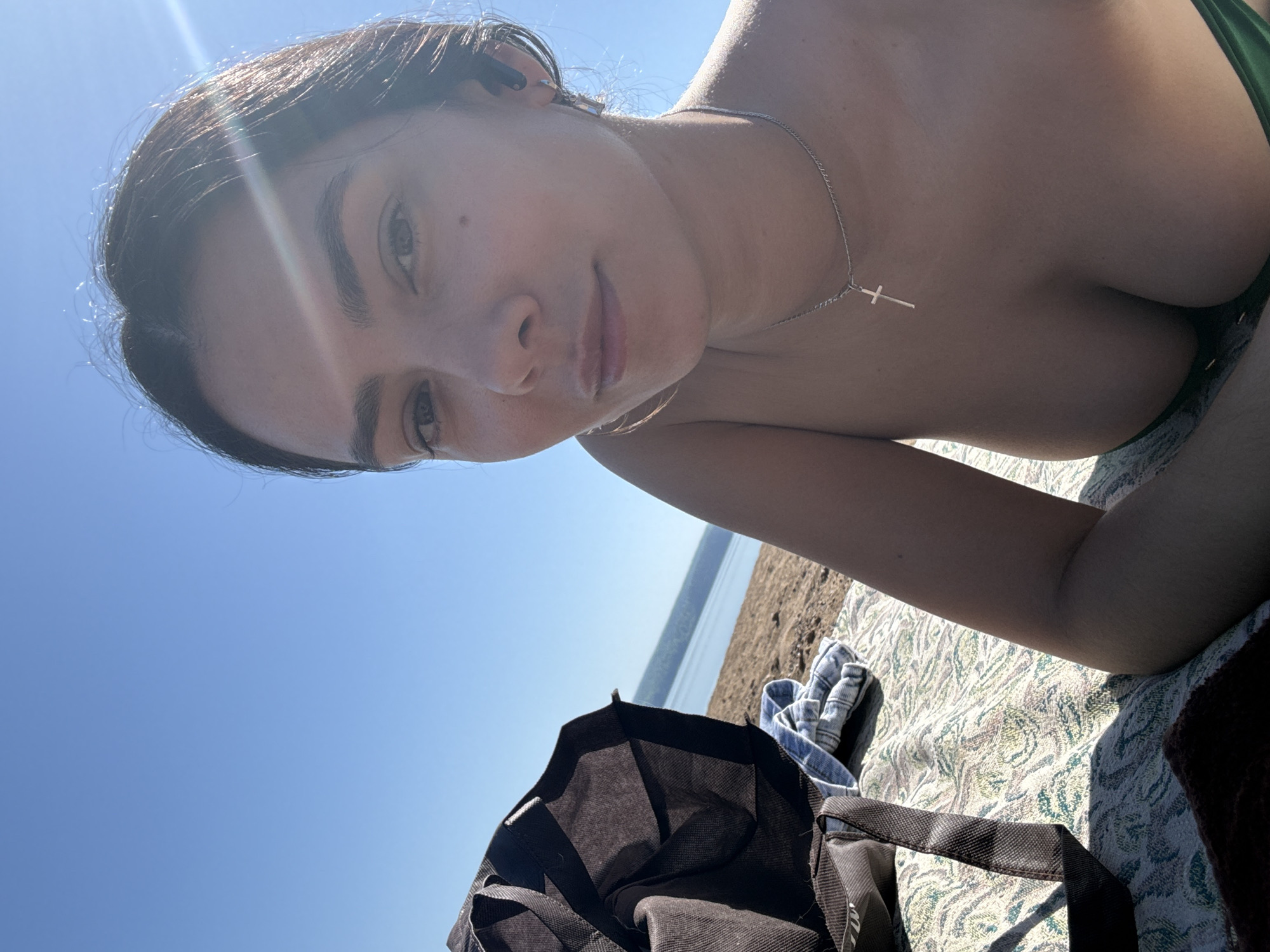 A young woman lies on a patterned blanket on a sandy beach under a clear blue sky. She is fair-skinned and wears a silver necklace with a cross pendant. Part of her green bikini top is visible. In the foreground, a black bag and a denim garment are on the blanket. The water and coastline of what is very likely the Kama River are visible in the background. This image was likely taken on a beach near the city of Chaikovsky, Russia.