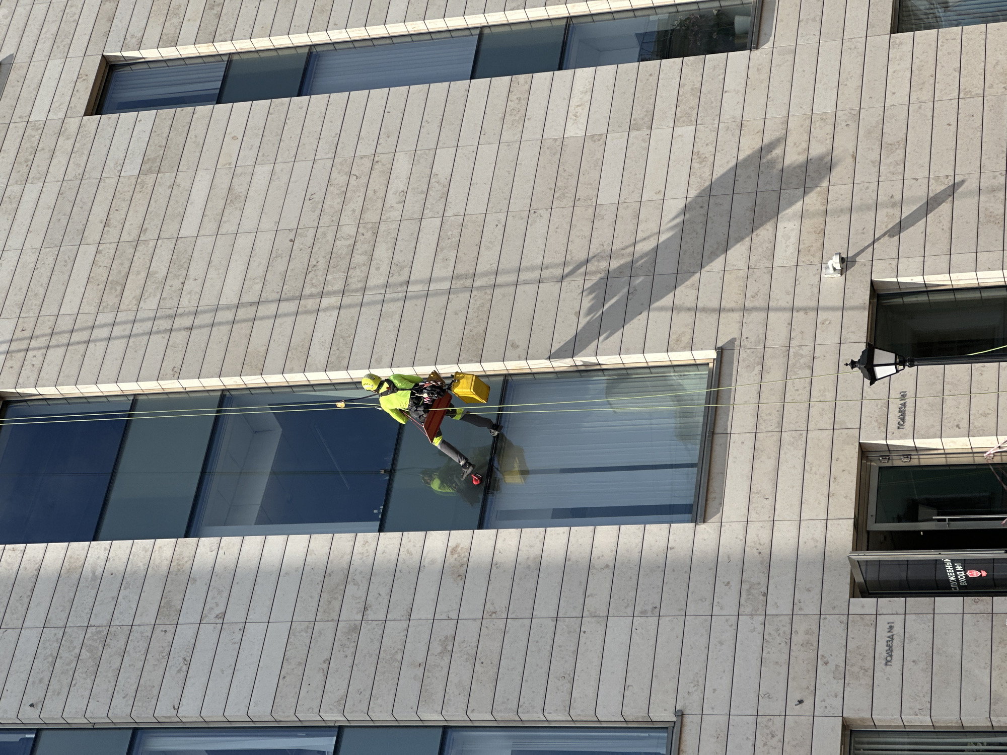 A window cleaner is suspended from the side of a building by two ropes, cleaning the windows. He wears a yellow helmet, a high-visibility yellow vest, gray pants, and red shoes. He is carrying a yellow bucket. The building has a light-colored stone facade with vertical lines. The windows are large and reflect the sky. The building appears to be located in the city of Saint Petersburg, Russia, as the inscription on the wall reads "ПОДЪЕЗД №1" which translates to "Entrance No. 1" in Russian. A street lamp is visible on the right side of the building.