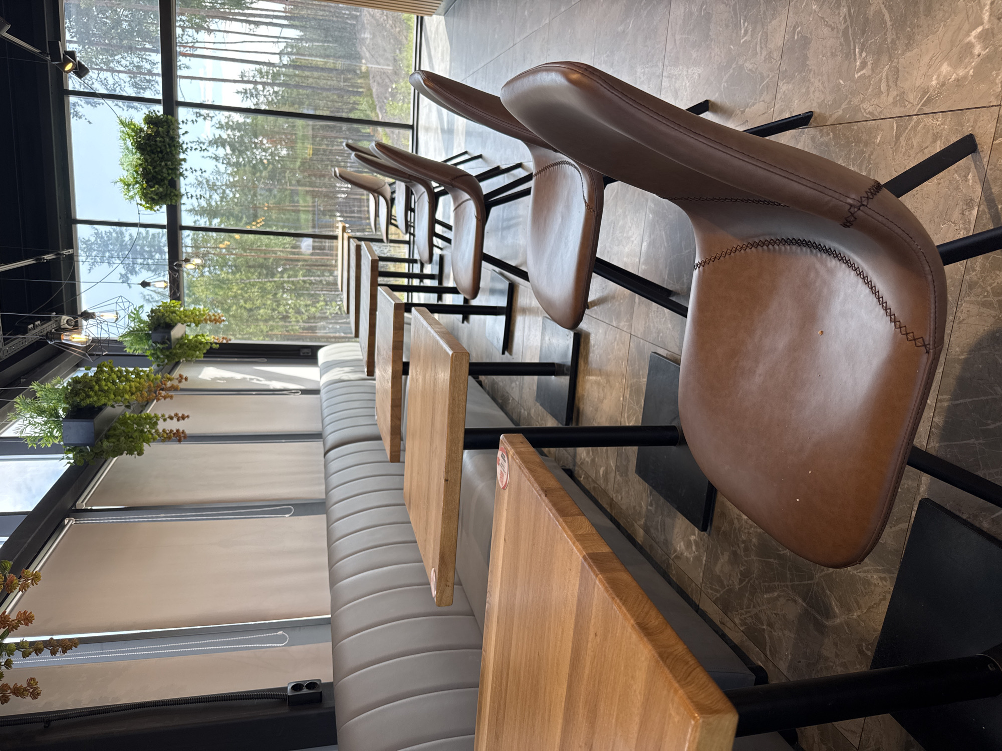 The image depicts the interior of a cafe with a row of empty tables and chairs. The tables are square with wooden tops and black metal bases. Several brown leather chairs with stitched detailing are arranged around the tables. In the background, a long upholstered banquette seating lines the wall. Large windows overlook a dense forest of pine trees under a clear sky. Hanging plants are visible both indoors and outdoors. The flooring is a dark, patterned tile. The scene appears to be in Peski, Russia, as indicated by the view of the surrounding forest.