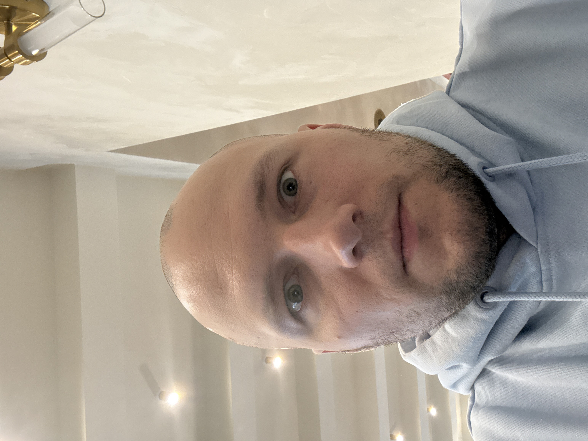 A man with a shaved head and a light-colored hoodie is facing the camera. The background appears to be an interior with light walls and multiple recessed lights. A brass and glass light fixture is visible in the upper left. The location in Krasnoyarsk, Russia is not discernable from the image.