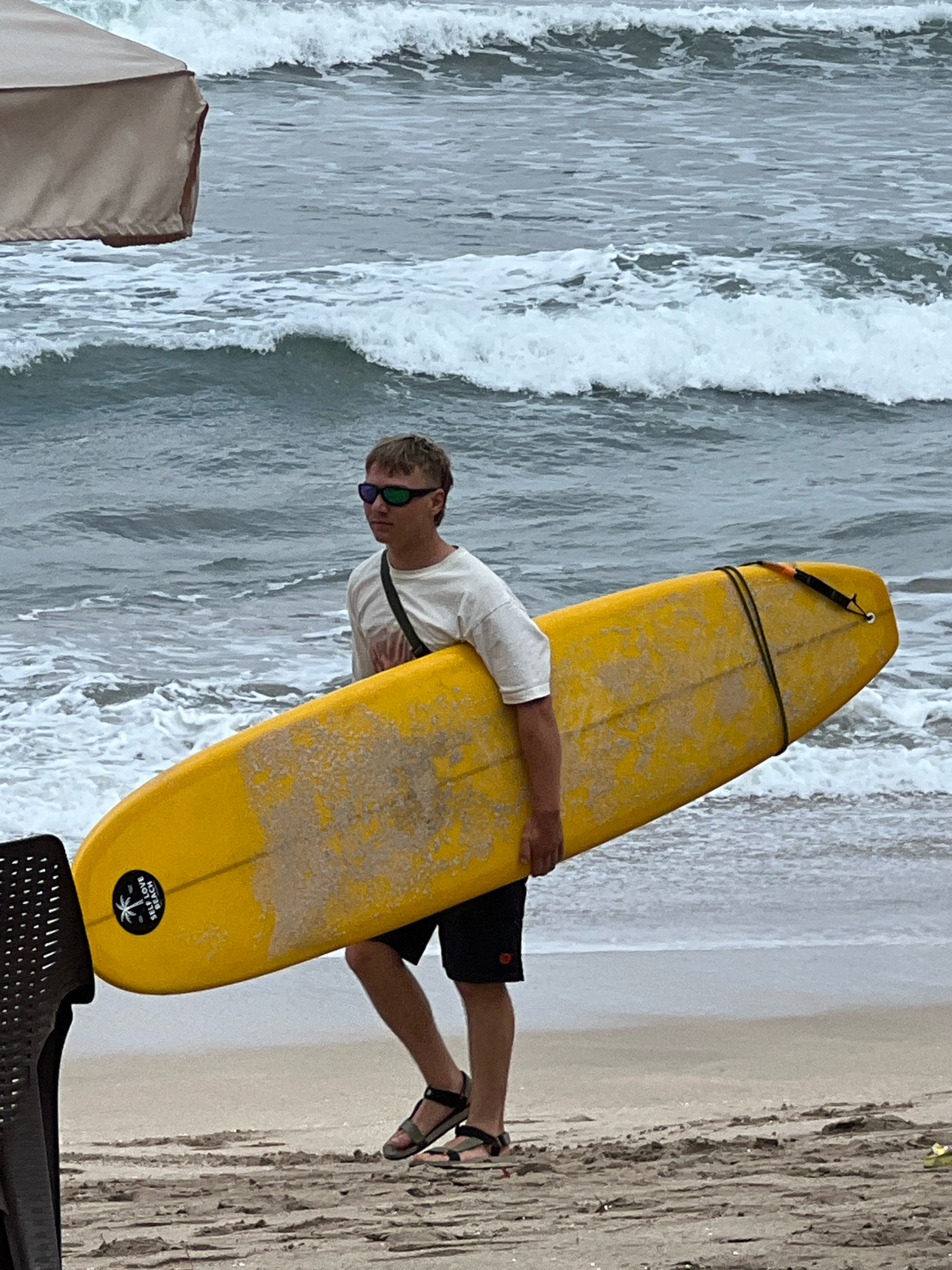 A young Caucasian male with short blonde hair, wearing sunglasses, a white t-shirt, black shorts, and sandals, is walking on a sandy beach in Kuta, Indonesia. He is carrying a yellow surfboard horizontally under his right arm. The surfboard has a black leash attached and a circular black sticker with a palm tree and the text "SELF LOVE BEACH" on the tail. The ocean is in the background with moderate waves breaking. To the left of the frame, a portion of a tan umbrella is visible. The sand on the beach is light brown and textured.