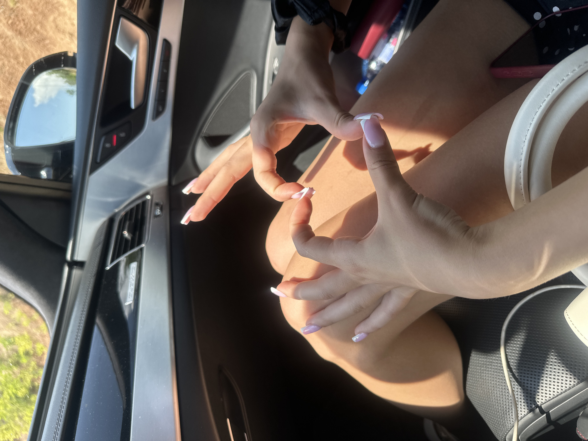 In Polikraishte, Bulgaria, a close-up shot captures two individuals' hands forming a heart shape inside a car. The hands have manicured nails with a French tip design. The interior of the car features black leather seats and silver trim. Sunlight illuminates the scene, casting shadows on the occupants' legs. Outside the car, greenery is visible through the window, suggesting a natural or rural setting.