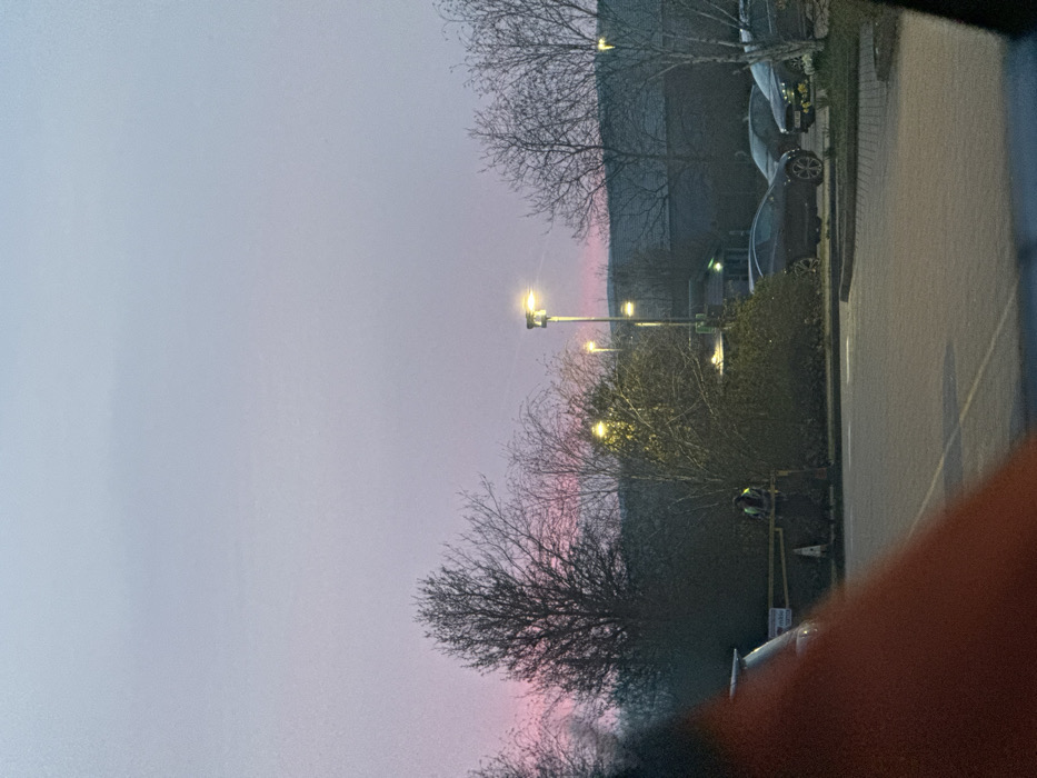 The image depicts an outdoor car park scene in Kettering, United Kingdom, under a low-light sky indicating either dawn or dusk. The upper sky is a light blue-grey, transitioning to a distinct pink-purple hue at the horizon.
In the lower-left foreground, a large, blurred reddish-orange object partially obstructs the view. The ground consists of an asphalt car park surface with white painted lines.
Mid-ground, a single individual stands upright to the left of the image center, positioned beside a horizontal yellow barrier and two orange/white traffic cones. The individual wears a high-visibility yellow-green vest with reflective stripes over dark clothing. Their orientation is generally towards the right of the frame.
Numerous deciduous trees with bare branches are visible across the mid-ground and background. Multiple streetlights with illuminated yellow-white lamps provide artificial light. In the background, a large, dark, low-rise building extends horizontally.
Several parked vehicles are present in the car park. A dark-colored hatchback/coupe is visible to the right of the individual, oriented left. To its right, a dark sedan and a lighter-colored vehicle are also parked. Smaller green structures are discernible near some of the streetlights.