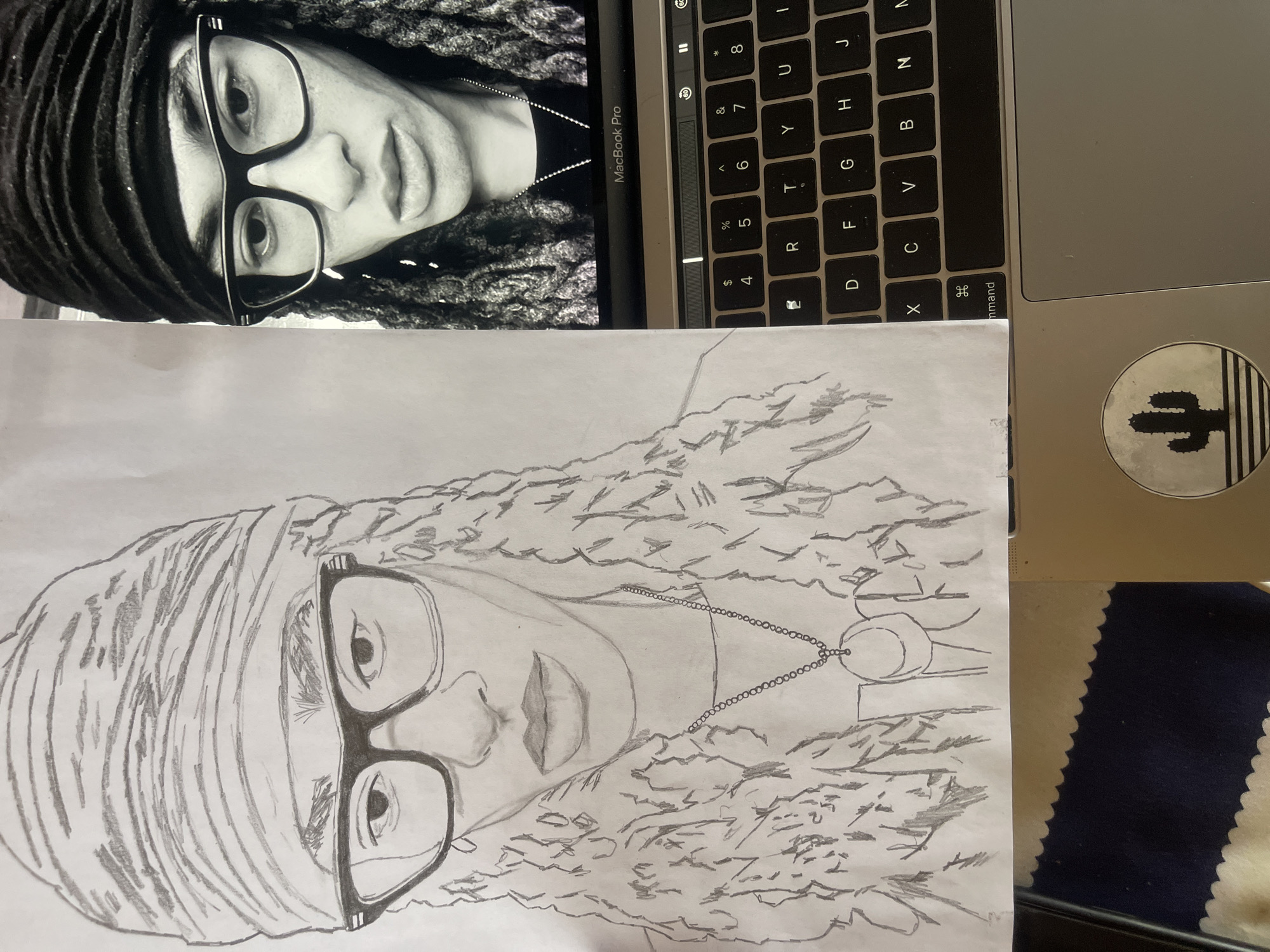 This image, captured in Jos, Nigeria, displays a pencil sketch and a digital photograph on a laptop screen, arranged on a flat surface. On the left, a hand-drawn pencil portrait on white paper depicts a person from the chest up, featuring a head wrap with horizontal stripes, thick-framed eyeglasses with light accents, and textured dreadlocks. A necklace with a circular pendant showing a crescent moon design is visible below the person's chin. To the right, a silver MacBook Pro laptop, with its lid partially open, displays a grayscale photograph of a similar individual on its screen. This digital image shows the person wearing a dark head wrap, matching dark-framed eyeglasses, and dark dreadlocks, with a subtle necklace chain. The laptop's keyboard and trackpad are visible in the foreground. A circular sticker featuring a black cactus silhouette against a striped background is affixed to the laptop's palm rest. The sketch paper partially overlaps the laptop's keyboard. A blue fabric or mat with a scalloped edge is visible beneath the sketch on the lower left.