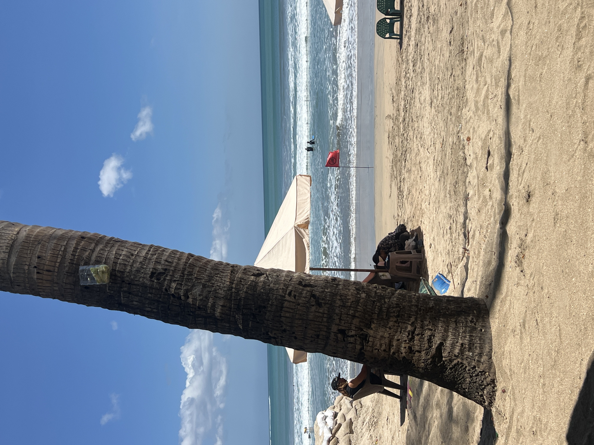 The image depicts a beach scene in Kuta, Indonesia. In the foreground, a large palm tree trunk dominates the left side of the frame, with a clear plastic bottle containing some light-colored items attached to its bark. The sand is light brown, showing footprints and some scattered debris. To the left of the palm tree, a person wearing a black shirt and a cap is seated in a dark chair, facing away from the camera and towards the ocean. Behind this person, a pile of sandbags is visible.
In the midground, a beige beach umbrella provides shade for two individuals. One person is seated on a stool, looking down at something on their lap, while another person is partially visible behind them, also seated. A red flag is planted in the sand in front of the umbrella. The ocean is blue with moderate waves breaking on the shore. Several people can be seen in the water, some appearing to be surfers. The horizon is clearly defined with a calm sea meeting a bright blue sky dotted with a few white clouds. In the far right of the midground, two green plastic chairs are positioned on the sand.