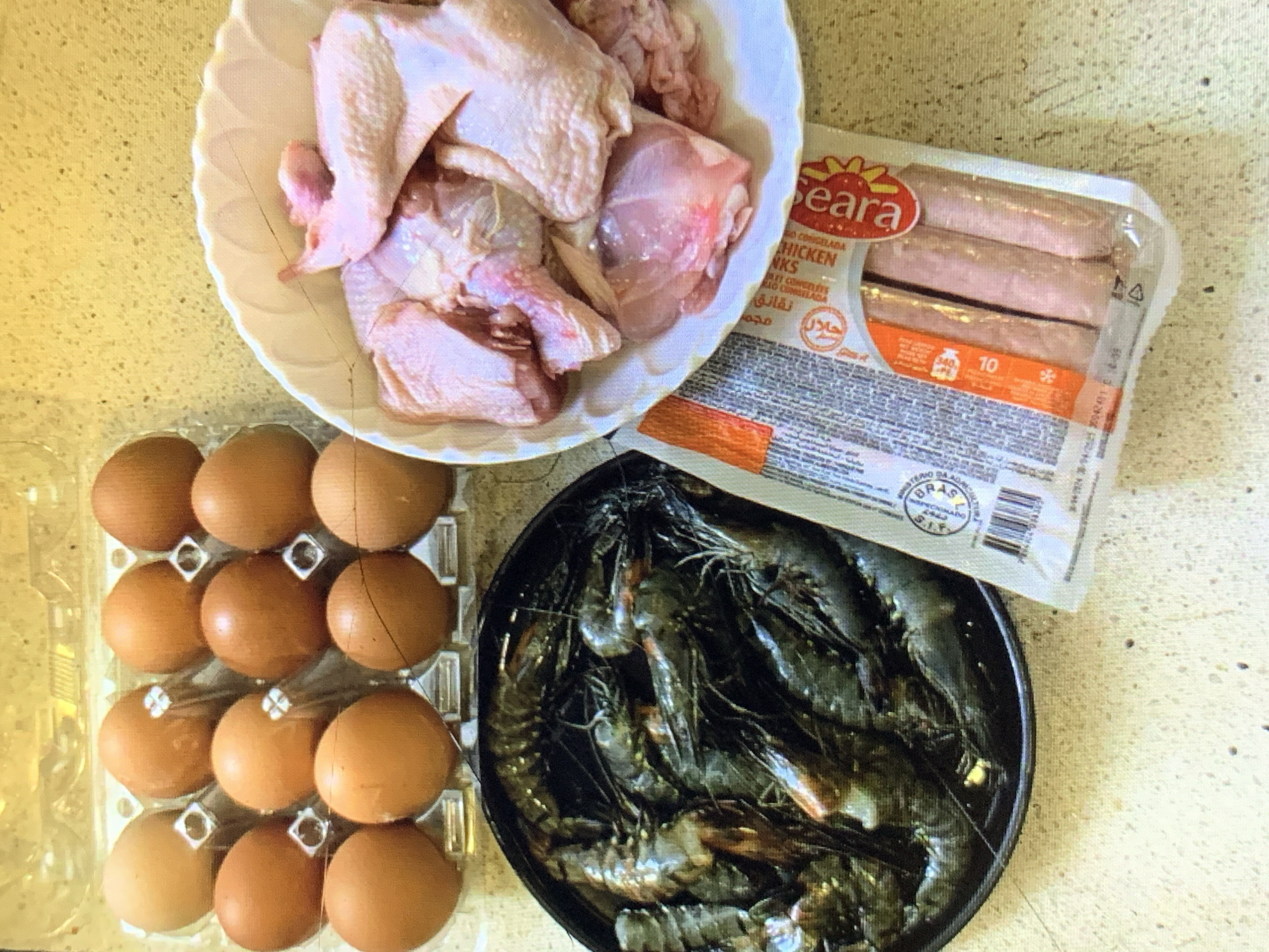 The image shows an overhead shot of ingredients placed on a speckled beige surface. The ingredients include a white bowl filled with raw chicken pieces, a package of "Seara" brand chicken franks, a plastic carton holding a dozen brown eggs, and a black bowl filled with raw prawns. The scene is likely in a kitchen or food preparation area. There is no indication that this image was taken in Rafin Par, Nigeria.