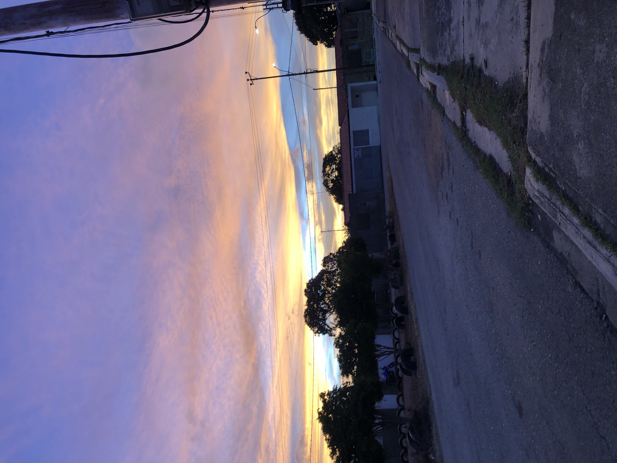 This image depicts a residential street scene in Altagracia, Venezuela, at either sunrise or sunset. The sky displays a gradient of colors, transitioning from deep purple and blue at the top to vibrant orange, yellow, and pink hues near the horizon, with scattered cloud formations visible throughout.
The foreground features a dark asphalt road with a visible texture, leading into the mid-ground. A concrete sidewalk and curb line the right side of the road, showing some cracks and interspersed patches of grass. A tall, light-colored utility pole with multiple dark electrical wires and an attached meter box is partially visible on the far right.
On the left side of the road, a dirt median is lined with numerous dark, horizontally placed tires. Behind this, a row of dark green, densely foliated trees partially obscures single-story residential structures with tiled roofs. A blue motorcycle is discernible parked in front of one of these houses on the left.
On the right side of the street, several single-story houses are visible, one featuring a white facade and a partially visible "Se Vende" (For Sale) sign on its garage door. Multiple utility poles and at least two streetlights are present along the road, one of which is illuminated. No individuals, animals, or active human interactions are depicted within the frame.