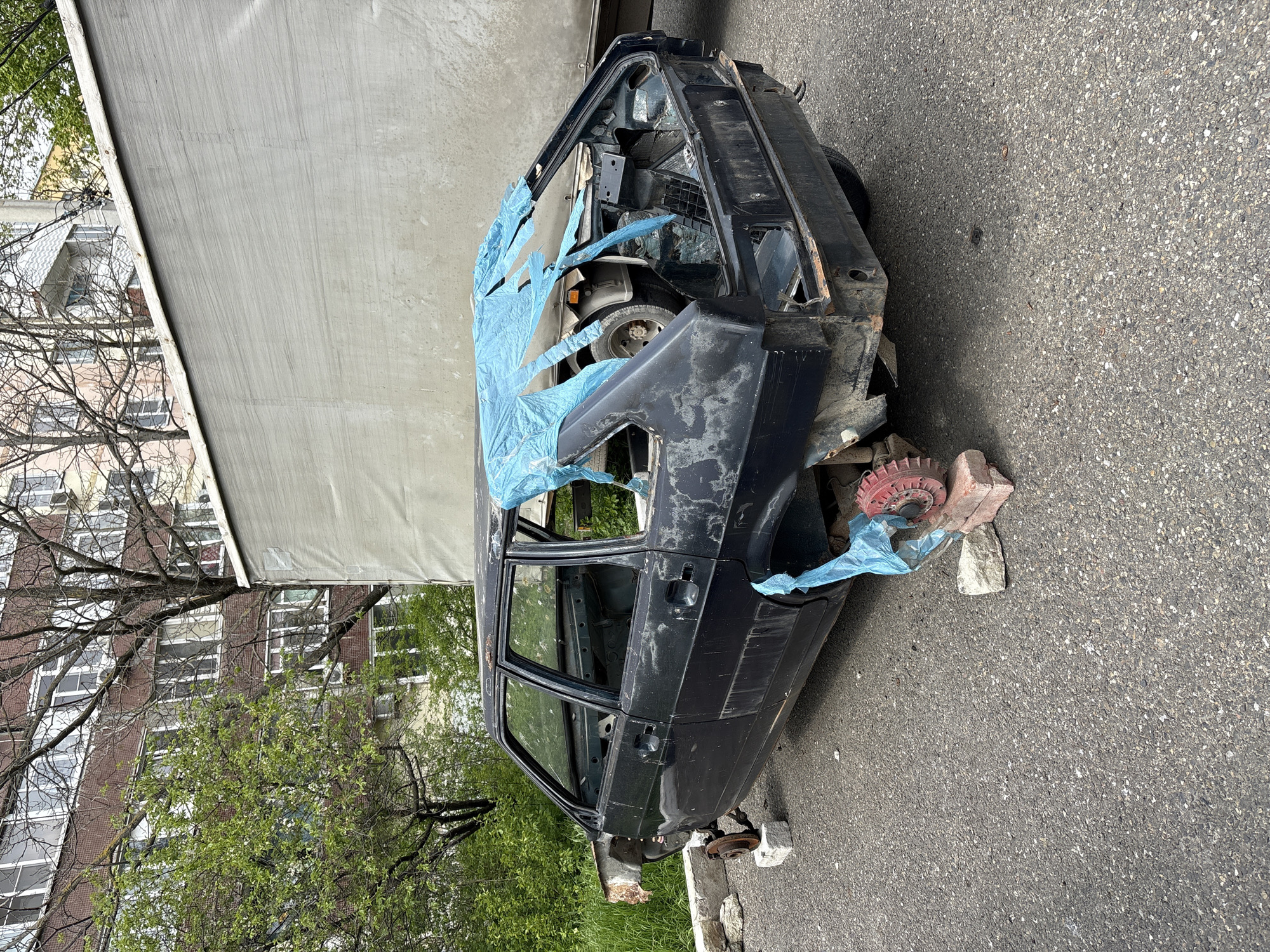 The image captures a derelict, heavily stripped black or dark blue car body, likely an older sedan, abandoned on an asphalt surface. The vehicle is missing its wheels, engine, and most interior fittings, indicating it has been dismantled for parts or left to decay. Blue plastic sheeting covers parts of the exposed interior and engine bay.
The car is precariously propped up; the front right side rests on bricks and a distinctive red, gear-like object (possibly a brake drum), while the rear is supported by other blocks. To the left and behind the car, a large, light-colored truck or trailer is partially visible. In the background, green trees with fresh spring foliage partially obscure a multi-story building with numerous windows, suggesting an urban or residential environment. The ground is paved with asphalt, transitioning to a grassy area on the left. The scene is illuminated by soft, even daylight, indicative of an overcast sky or diffuse lighting. No discernible text is visible. The general appearance of the vehicle and surroundings is consistent with an urban setting in Gelendzhik, Russia.