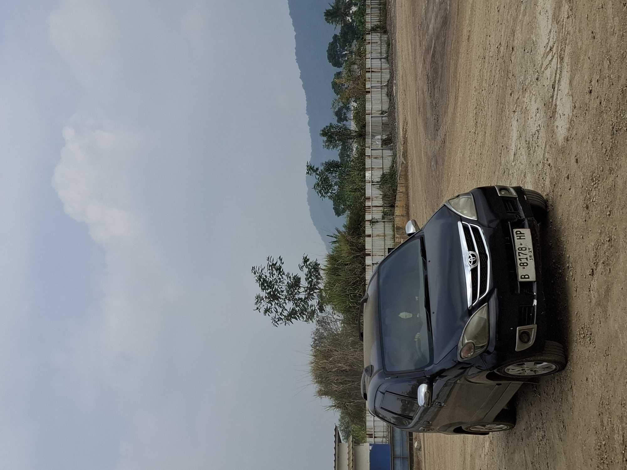 A black Toyota Avanza, license plate B 8178 HP, is parked on a dusty, uneven dirt lot. The vehicle is facing slightly to the right, with its headlights on. The car is a multi-purpose vehicle, with roof rails and a silver-colored grille. The background shows a chain-link fence with corrugated metal panels, beyond which are trees and a hazy mountain range. The sky is overcast with visible clouds. Part of a light blue and white building is visible to the far left. This scene is located in Kutasirna, Indonesia.