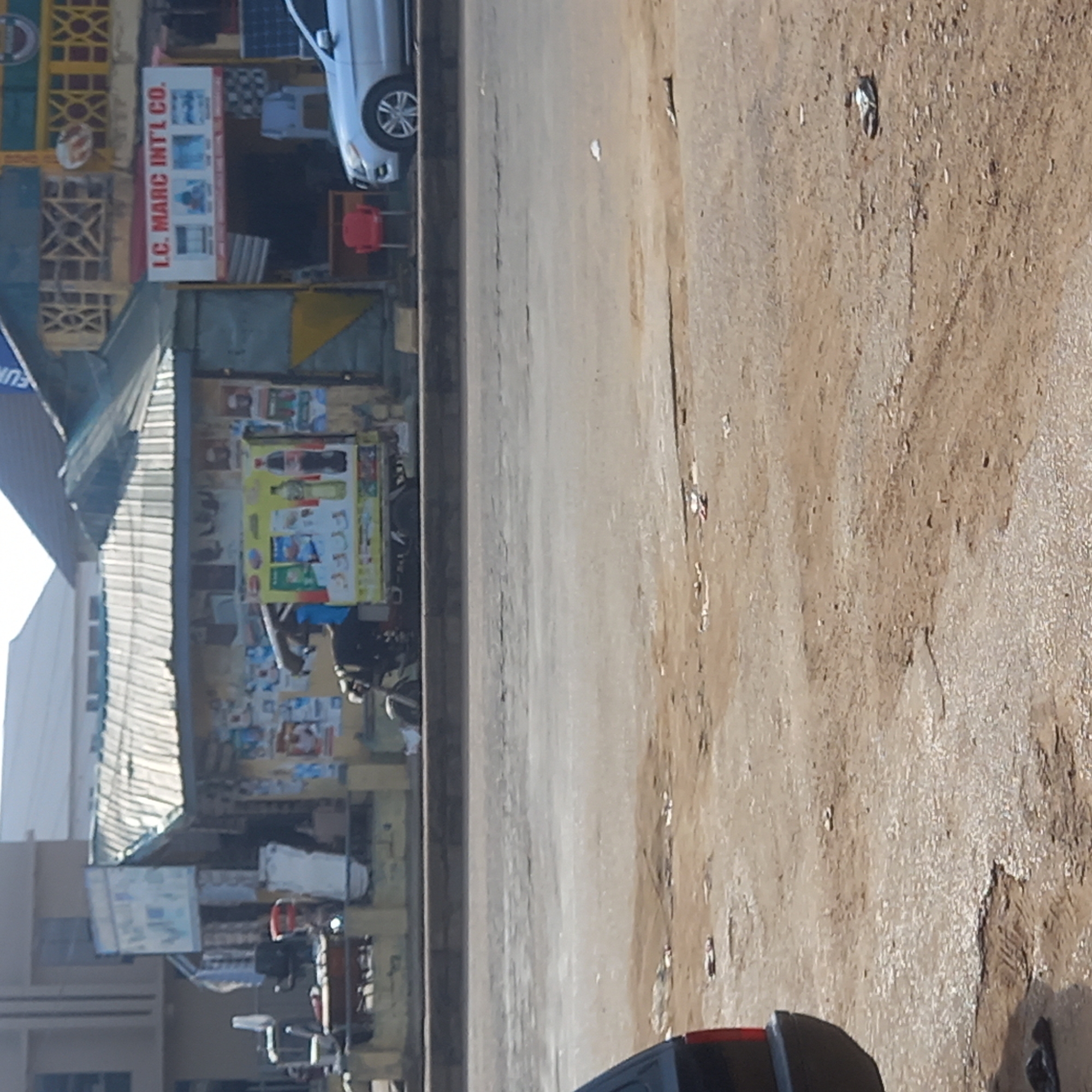 The image depicts a street scene in Dadi Riba, Nigeria. On the left side of the image, a building with shops is visible, featuring advertisements and signage, including "L.C. MARC INT'L CO." A vehicle is parked near the building. The street itself is wide and appears to be paved, with visible wear and debris. A motorcycle is parked in front of one of the shops. The overall impression is of a typical commercial street in a Nigerian city.