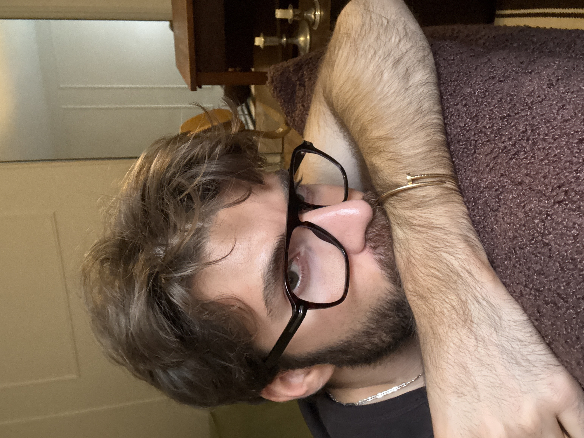 A close-up, slightly angled shot depicts a man with light brown hair and a trimmed beard, wearing black-rimmed glasses. He rests his head on a brown textured cushion or pillow. His left forearm, adorned with a gold bracelet resembling a nail, is positioned across his face. A silver chain necklace is visible around his neck, and he is wearing a dark-colored shirt. The background includes a light-colored wall and a wooden object, possibly a piece of furniture, with decorative candlesticks on top. There is no contextual information to determine the location as Montreal, Canada.