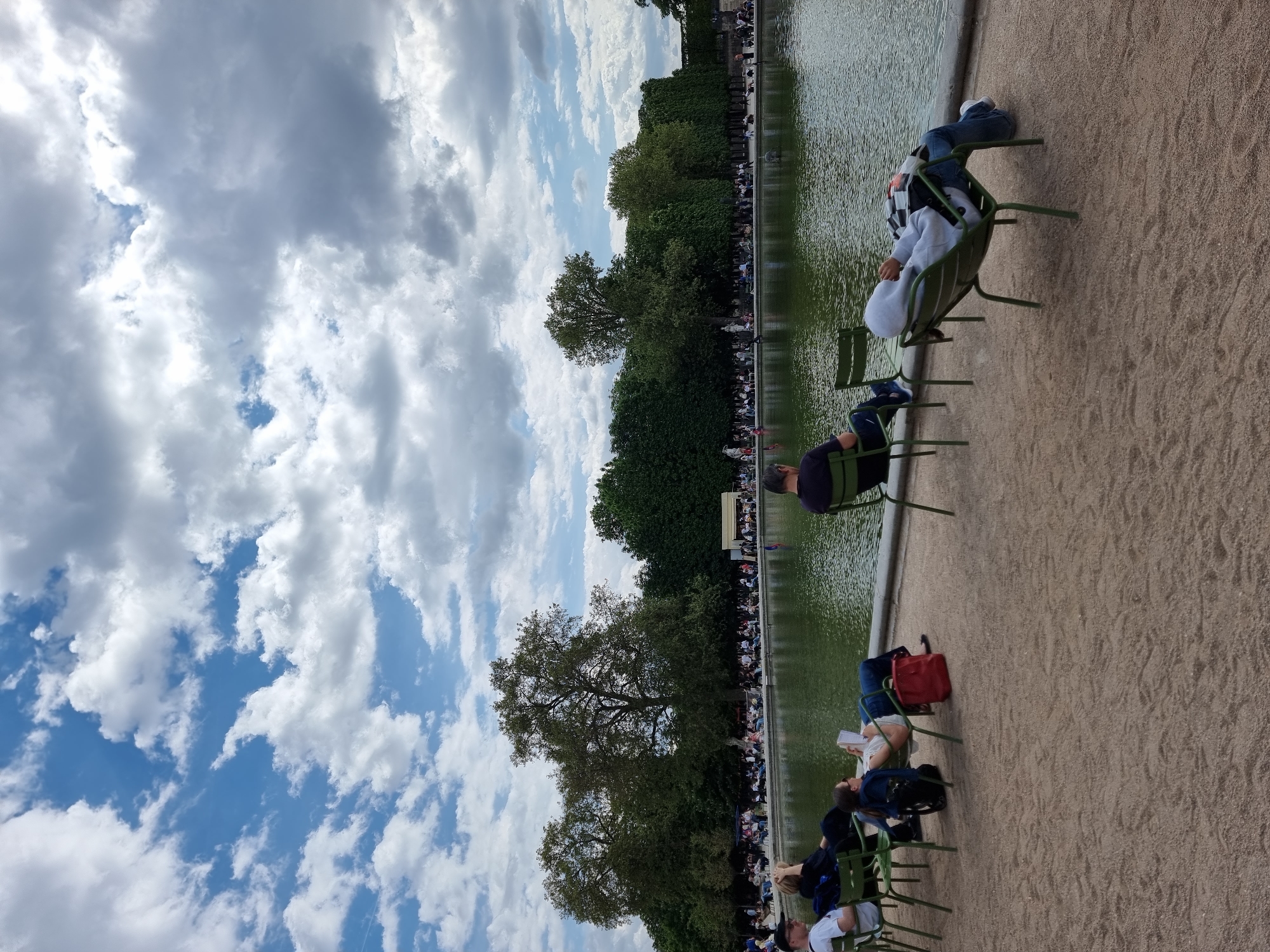 This outdoor image captures a scene at the Jardin des Tuileries in Paris, France. People sit in green metal chairs along the edge of a large pond. A line of trees and other people are visible on the opposite side of the pond. The sky is filled with clouds and patches of blue. The ground is a light brown color. The people are relaxing and enjoying the scenery.