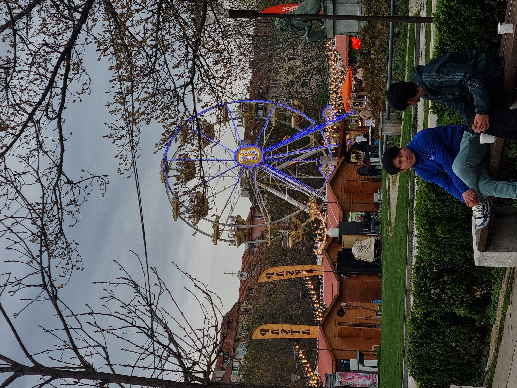 The image depicts a scene in Sofia, Bulgaria, likely at the Christmas market. A large Ferris wheel with blue lights and carriages is a central feature. Several wooden stalls, decorated with lights, are visible below the Ferris wheel. Bare trees are in the foreground, partially obscuring the view. Two young men are sitting on a bench on the right side of the image. One is wearing a blue jacket and green pants, and the other is wearing a dark jacket. Buildings are in the background. The sky is overcast.