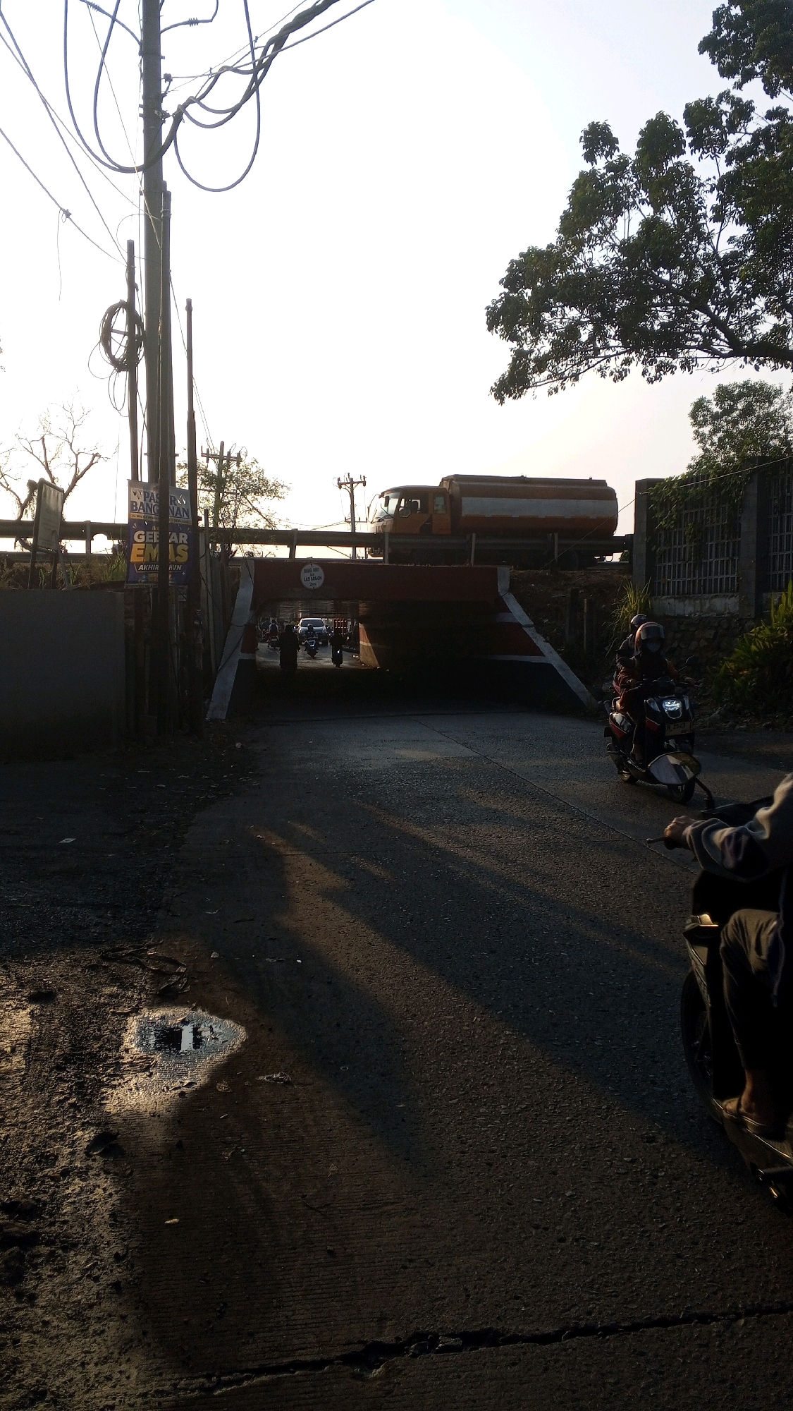 This image depicts a street scene in Semarang, Indonesia, likely during the late afternoon given the long shadows cast by the setting sun. A road runs towards a railroad overpass, under which several vehicles are passing. A tanker truck is visible on the tracks above. On the right side of the road, two motorcyclists are riding towards the viewer, their faces obscured by helmets. The road surface is asphalt, with some patches of wear and tear, and a small puddle of water reflects the sky on the left side of the foreground. To the left of the road, a utility pole with numerous wires stands prominently, with a sign attached to it advertising "PASAR BANGUNAN". Vegetation, including trees, can be seen on the right side of the overpass. The overall atmosphere is one of everyday urban life in Semarang.